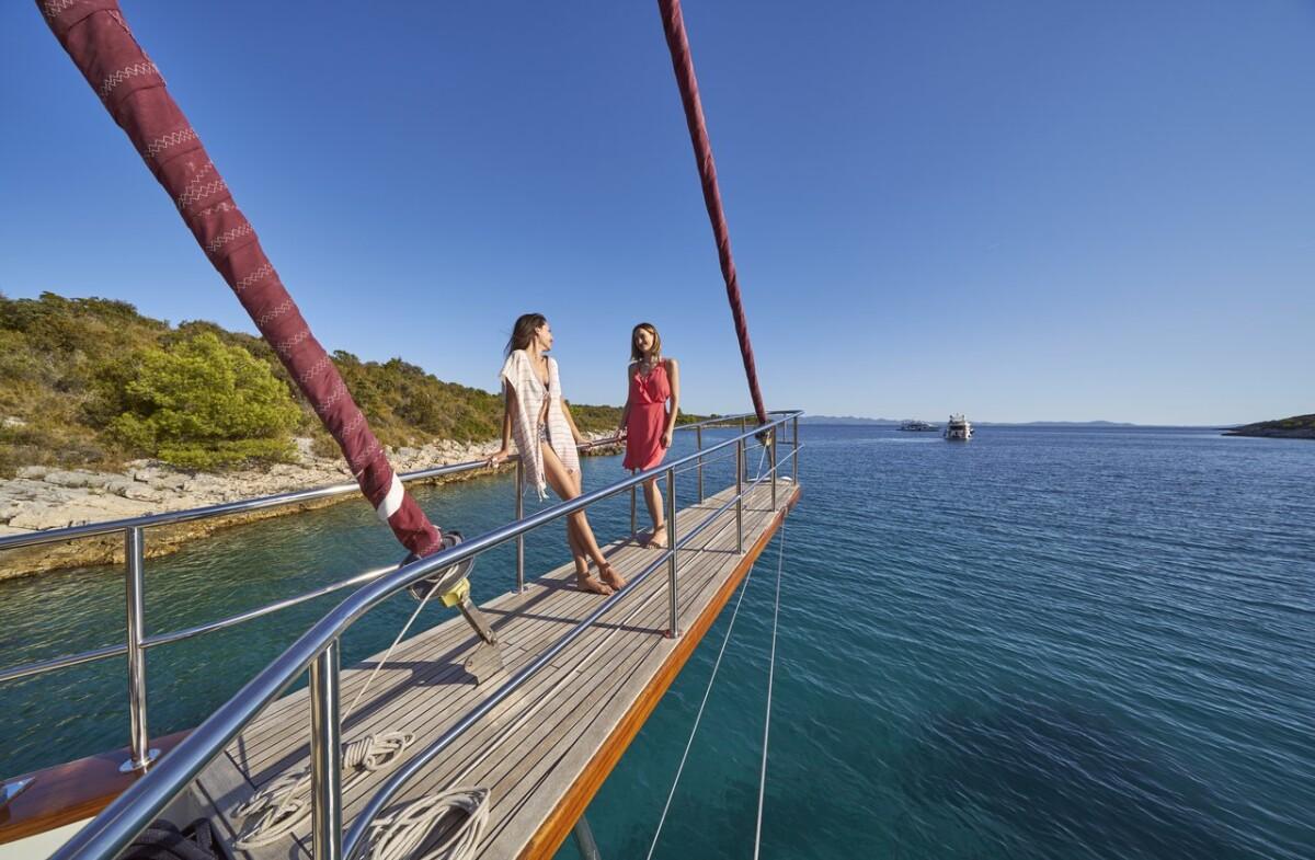 Two women standing on gulet bow deck with steel railings overlooking turquoise bay