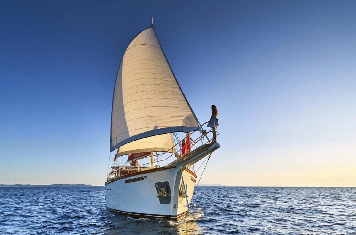 White sailing yacht with cream mainsail and person on bowsprit sailing on blue water
