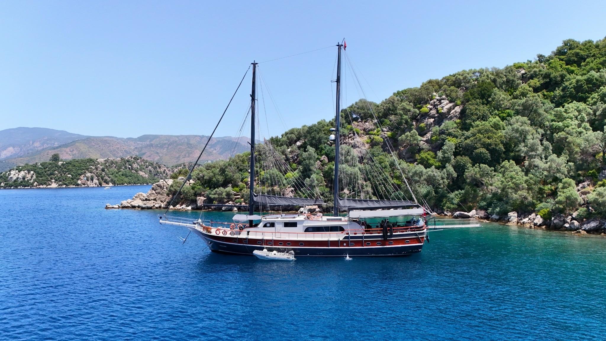 traditional wooden gulet with two masts anchored in crystal clear turquoise bay surrounded by forested hills