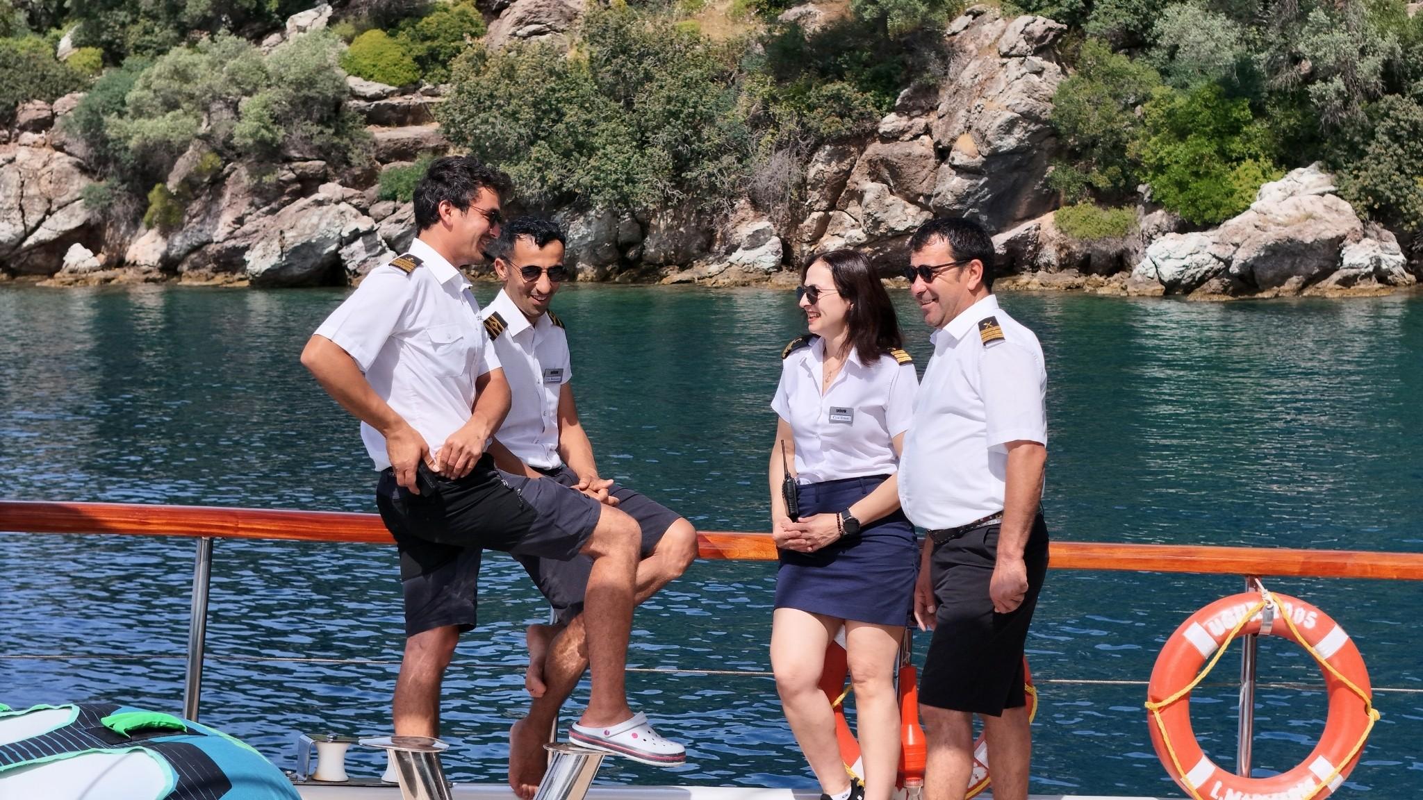 Four yacht crew members in white uniforms relaxing on deck with turquoise water and rocky coastline