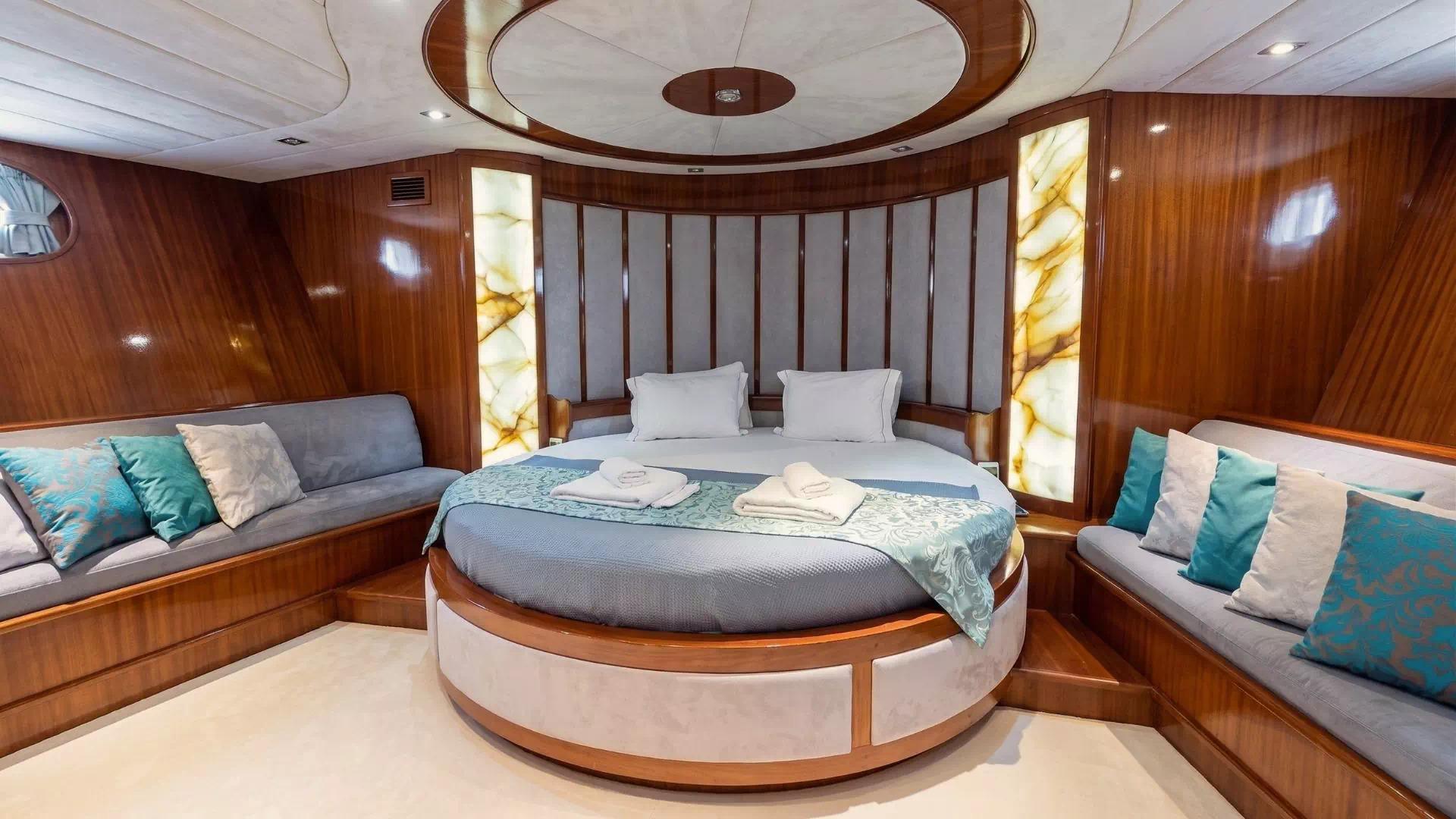 Master cabin featuring circular bed, curved headboard, built-in seating with teal cushions, and polished wood finishes