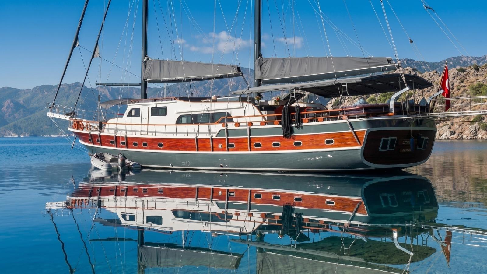 Traditional Turkish gulet with varnished mahogany hull and grey topsides anchored in glassy calm bay with mountain backdrop