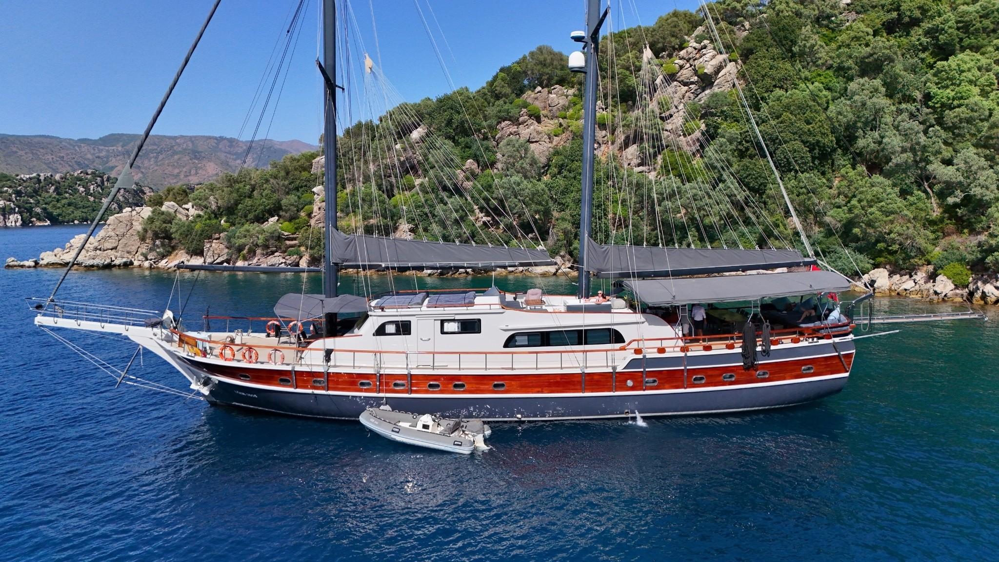 Luxury gulet with dark hull and varnished teak strake anchored in turquoise cove with rocky forested coastline