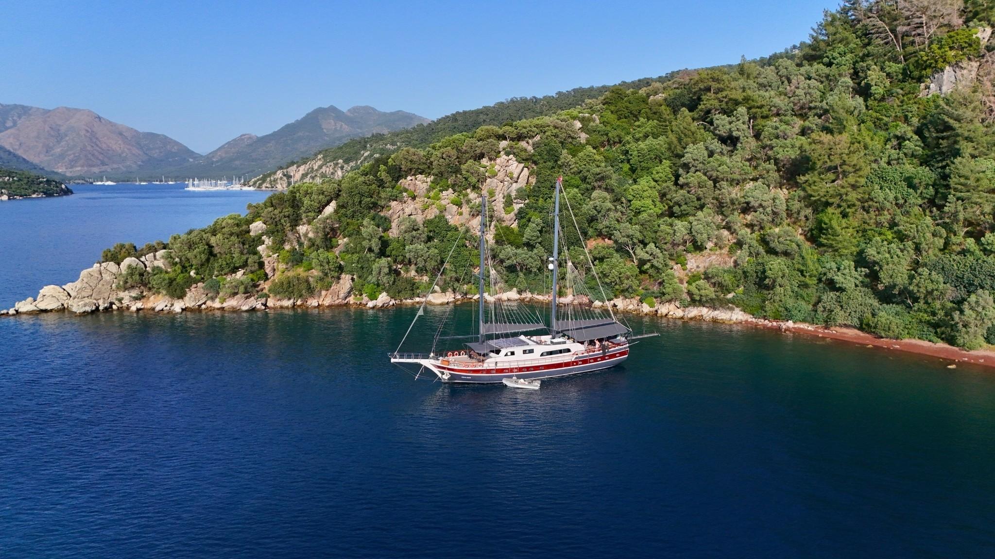 Aerial drone view of red-hulled gulet anchored in calm turquoise cove with forested rocky coastline