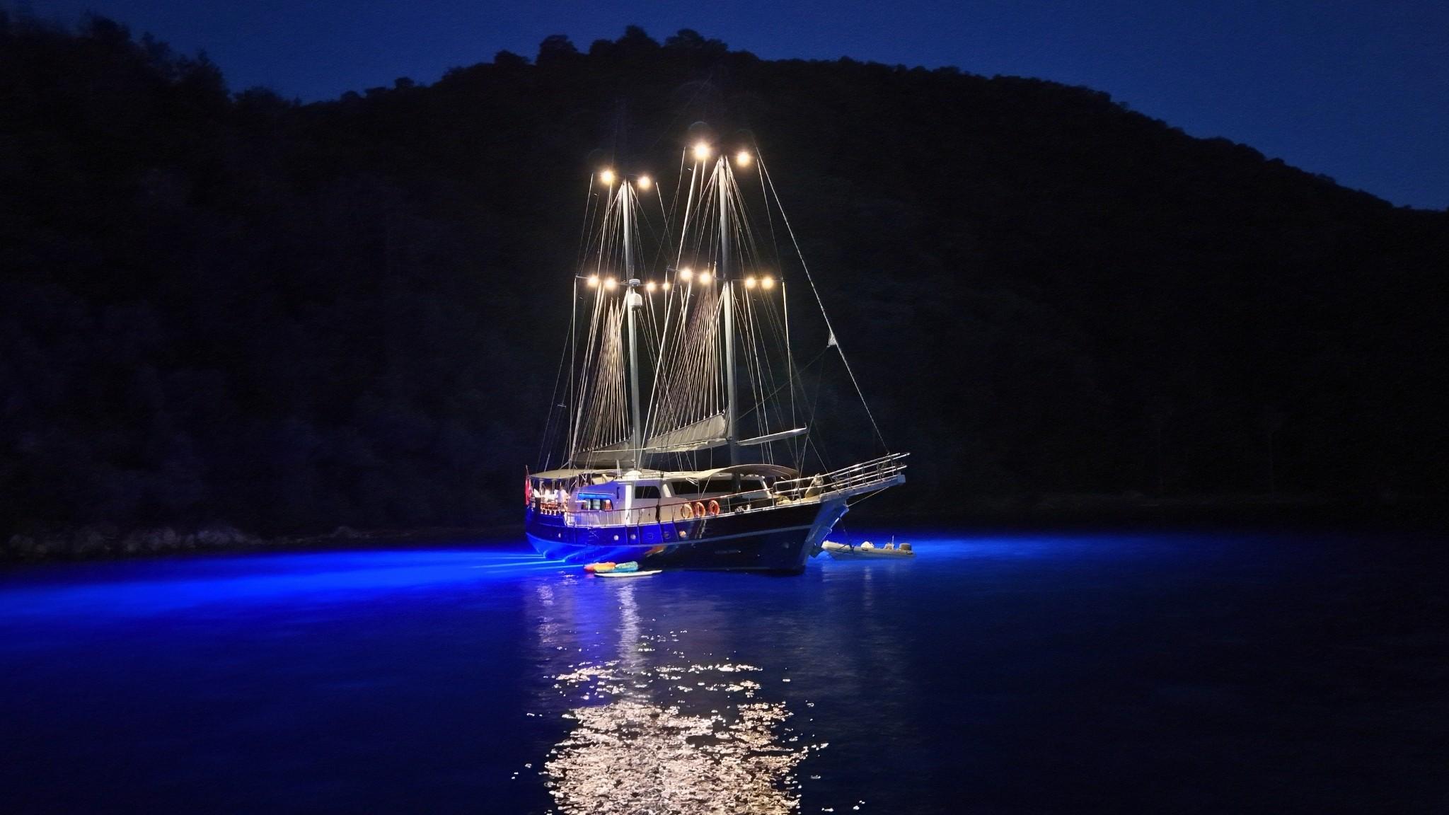 luxury gulet anchored in calm bay at night with glowing blue underwater hull lights and illuminated masts
