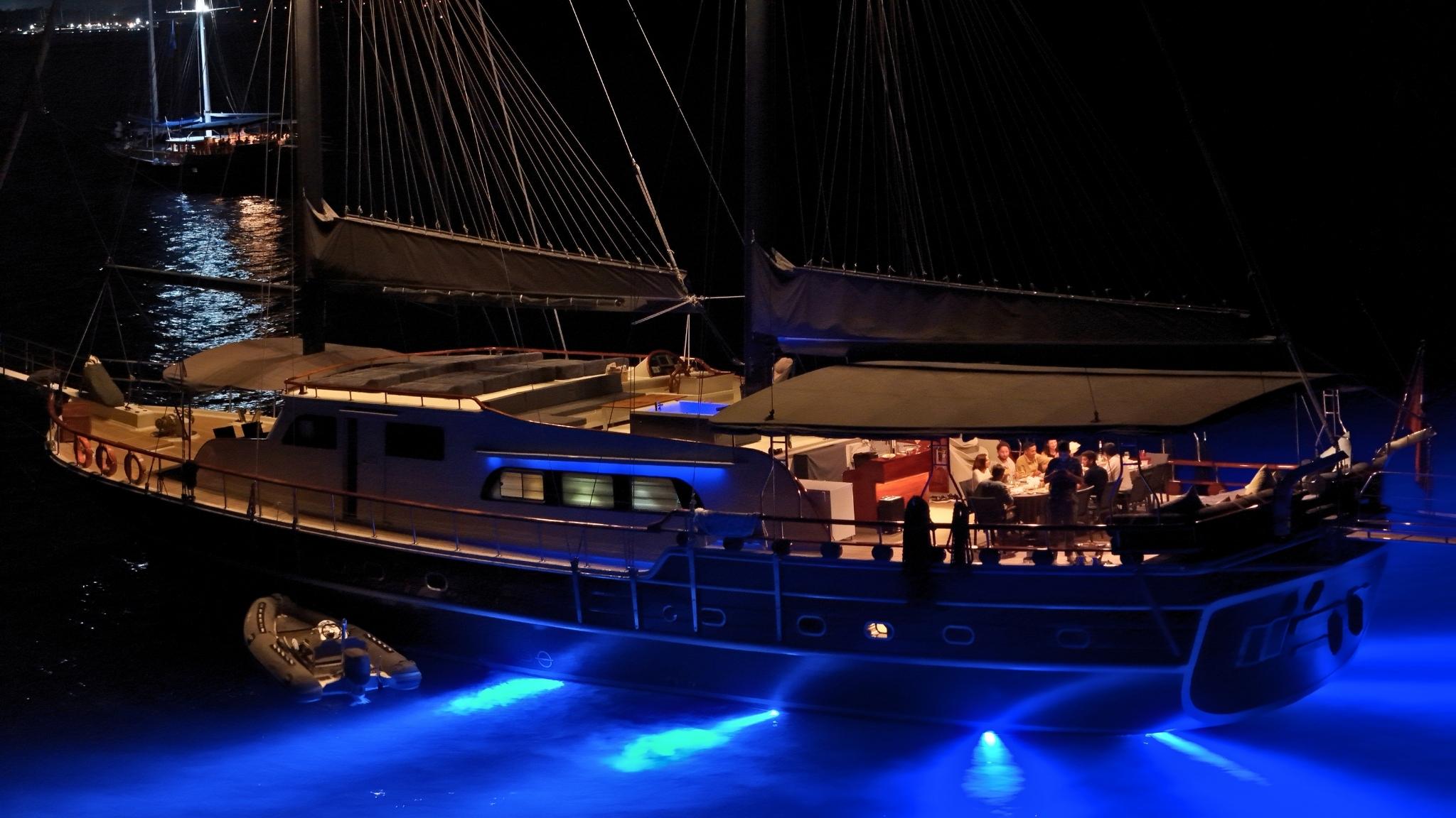 Large sailing gulet moored at night with vivid blue underwater hull lights illuminating the sea, guests dining on aft deck, tender alongside