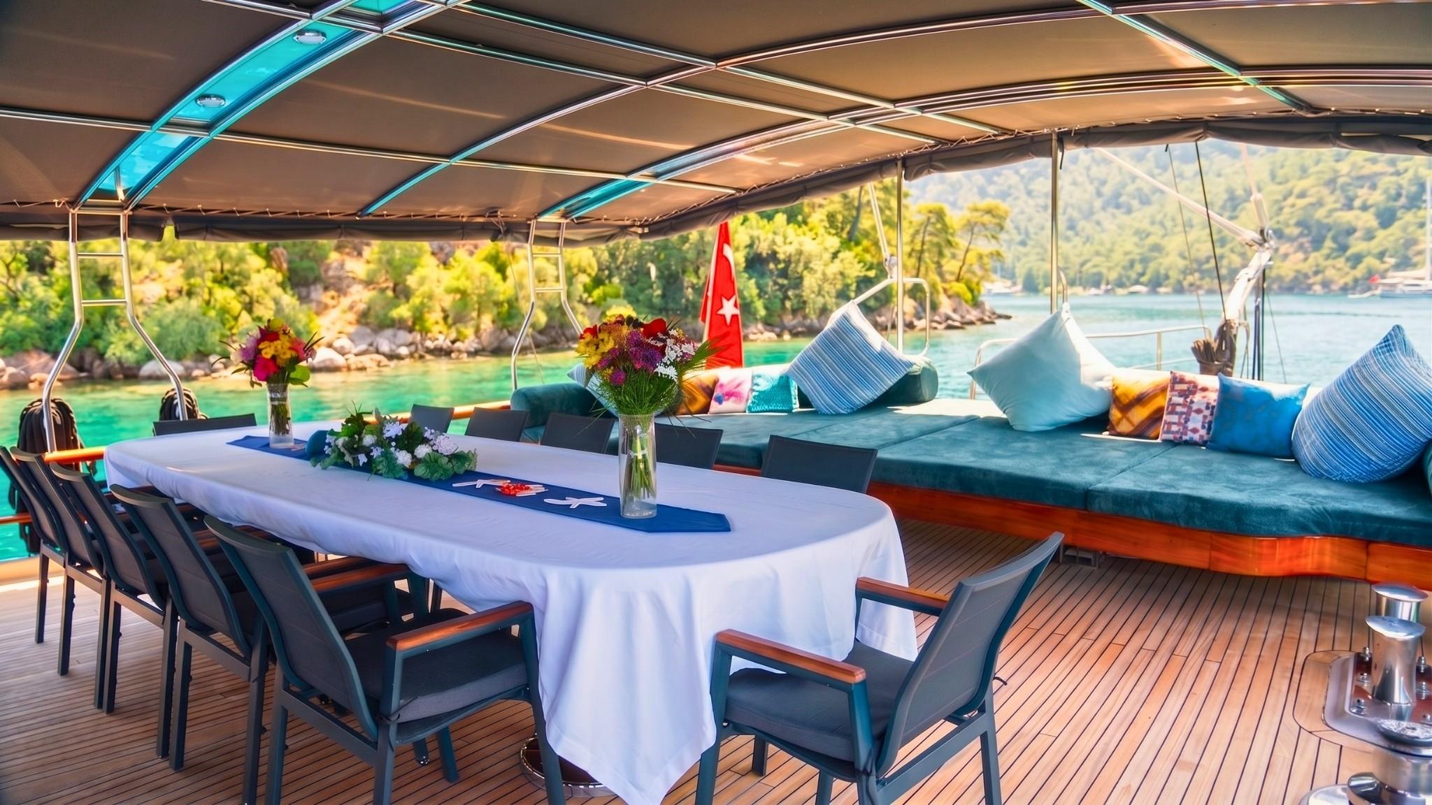 Gulet aft deck dining table set with white linen and flowers, teal cushioned bench seating alongside, anchored in turquoise bay