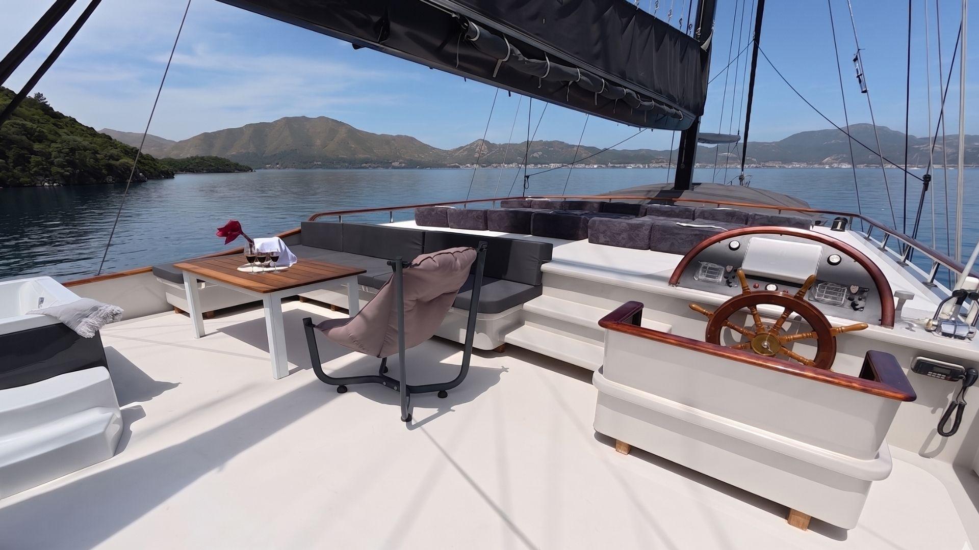 luxury gulet cockpit featuring wooden helm wheel, grey upholstered seating, teak dining table with two red wine glasses, and mountain anchorage backdrop
