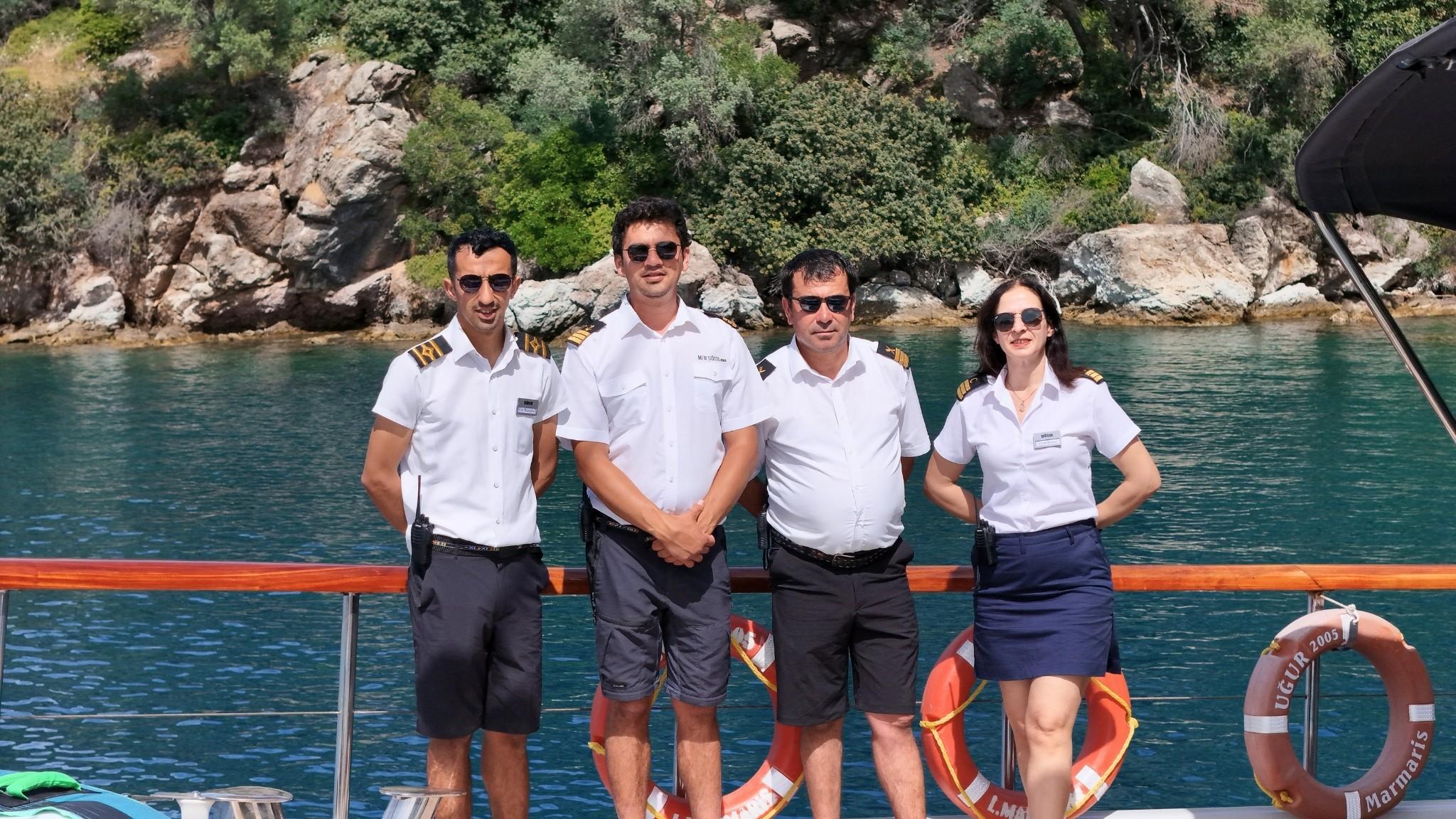 Four uniformed crew members standing on the deck of gulet Ugur 2005 registered in Marmaris with rocky coastline behind