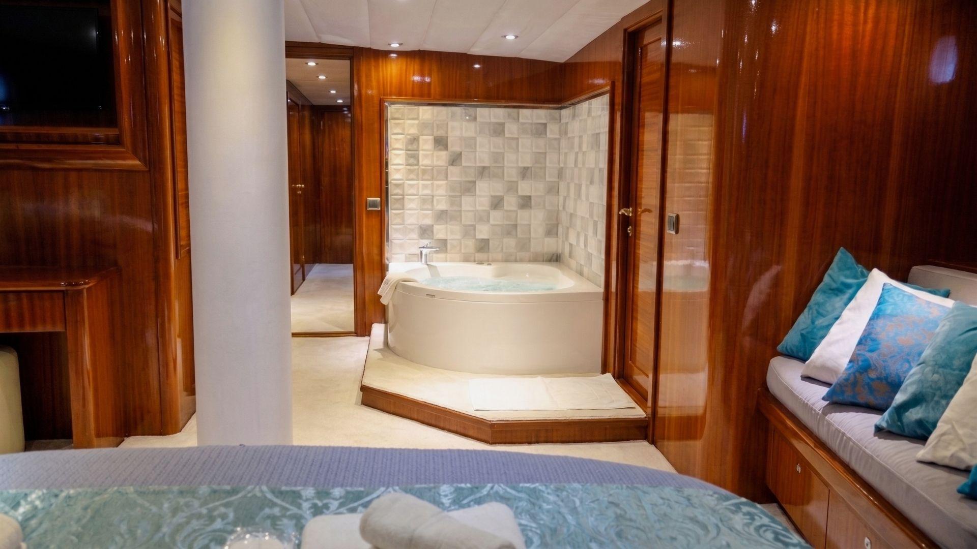 Master cabin en-suite bathroom with corner jacuzzi bathtub set on raised wooden platform against tiled wall, with seating area and flat-screen TV visible