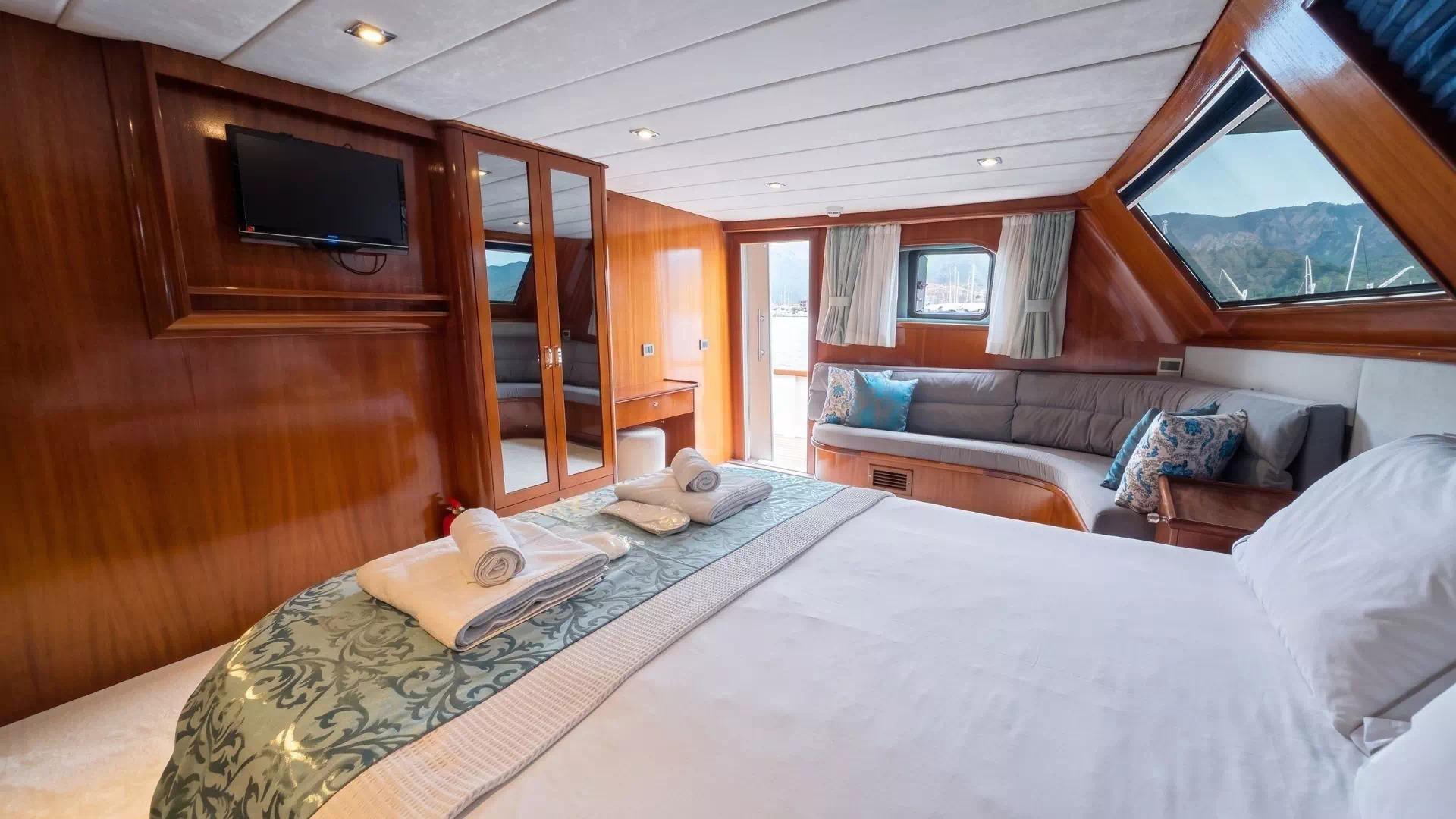 Gulet master cabin featuring king bed with decorative runner, built-in sofa seating with blue cushions, mirrored wardrobe, wall-mounted TV and large panoramic windows overlooking mountains