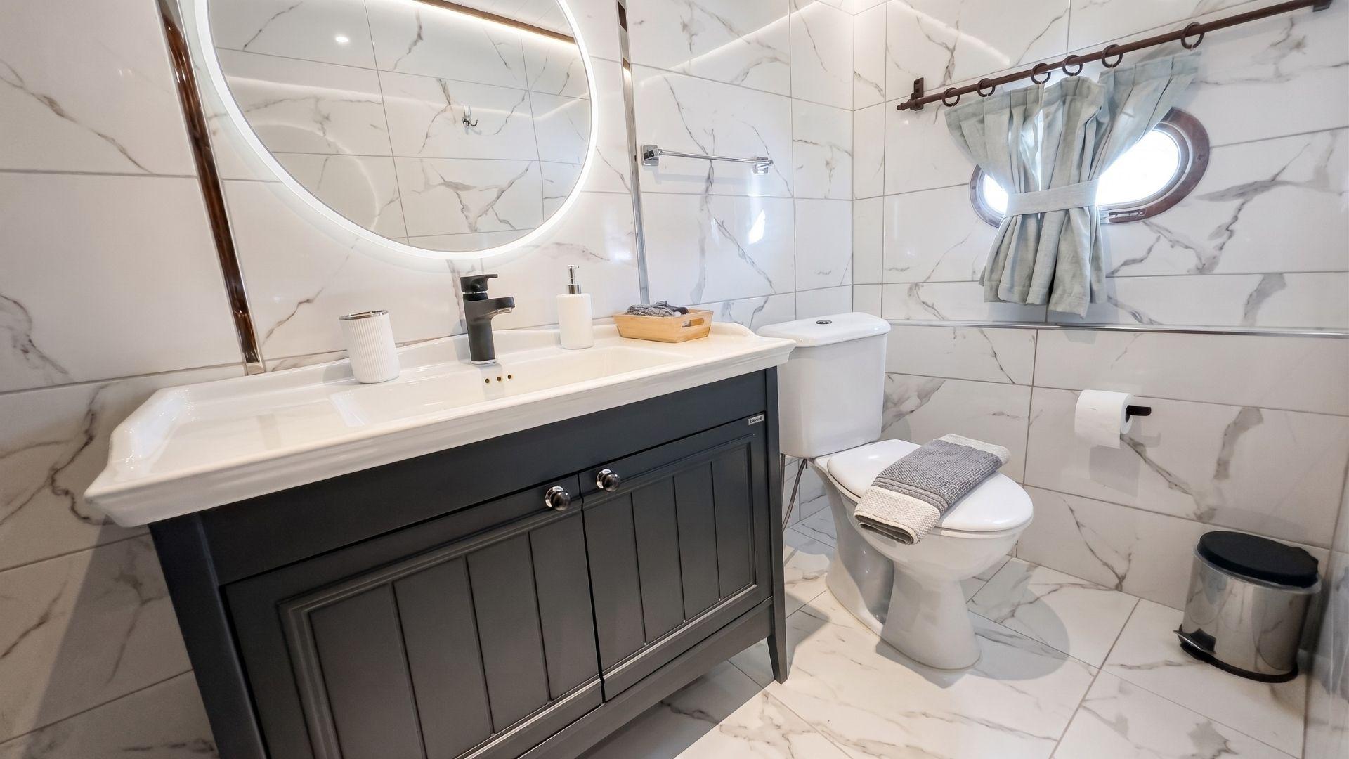 Yacht en-suite bathroom with dark vanity unit, backlit round mirror, white marble-effect tiles and oval porthole window