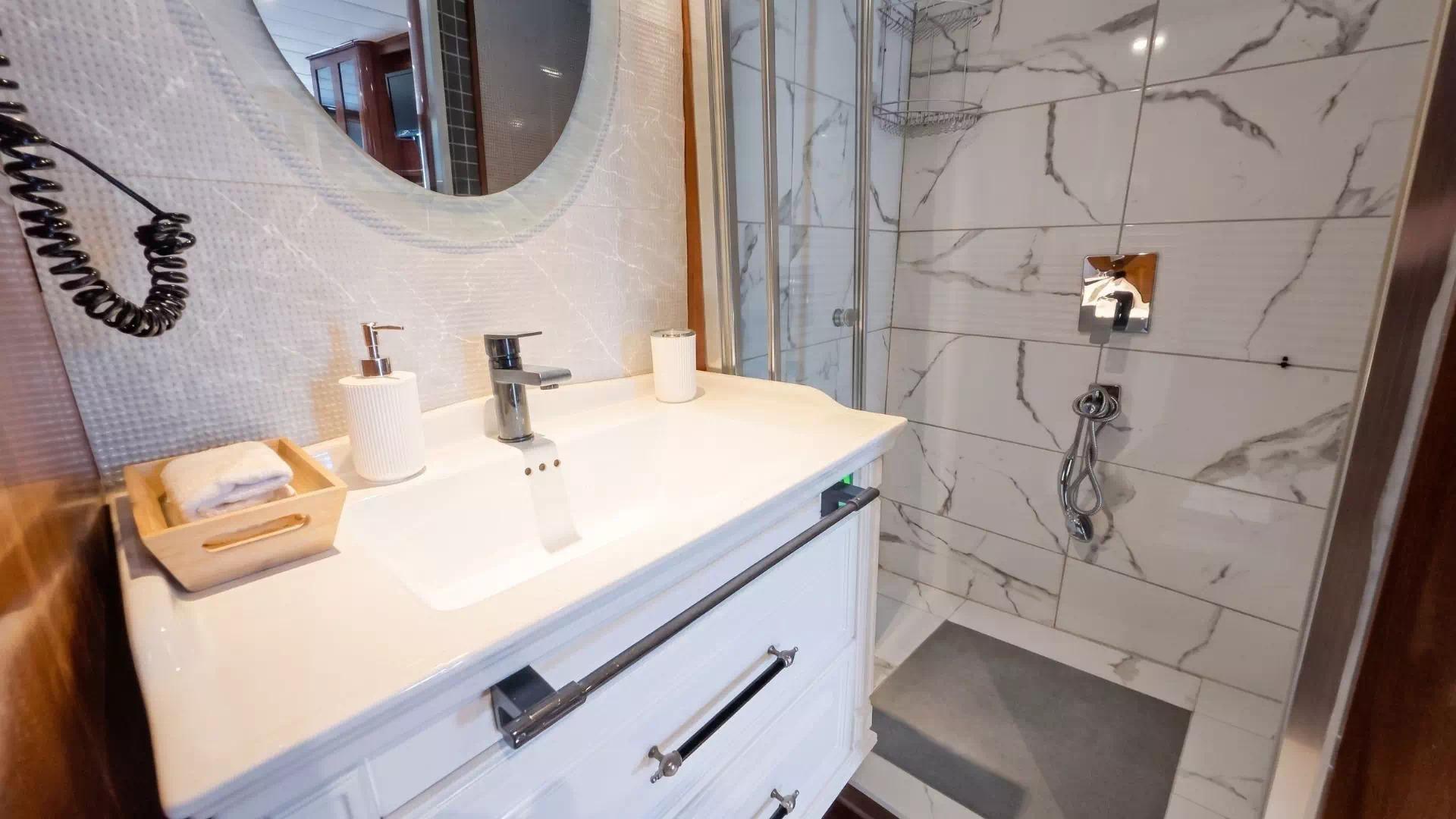 Yacht cabin en-suite bathroom with white vanity unit, round mirror, and marble-tiled walk-in shower