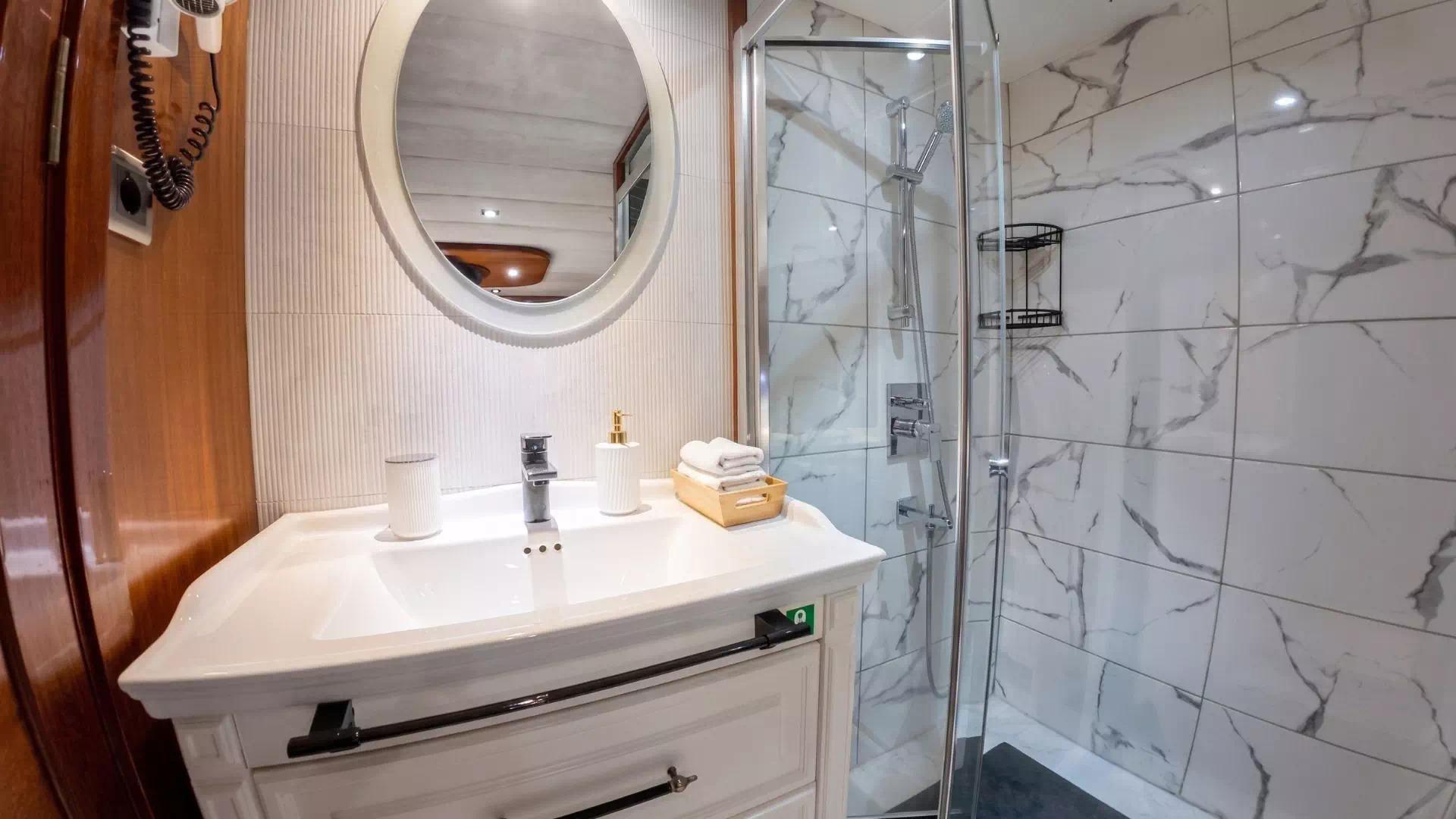 Luxury gulet cabin en-suite bathroom with marble-effect tiled walk-in shower, white vanity unit, round illuminated mirror and chrome fittings