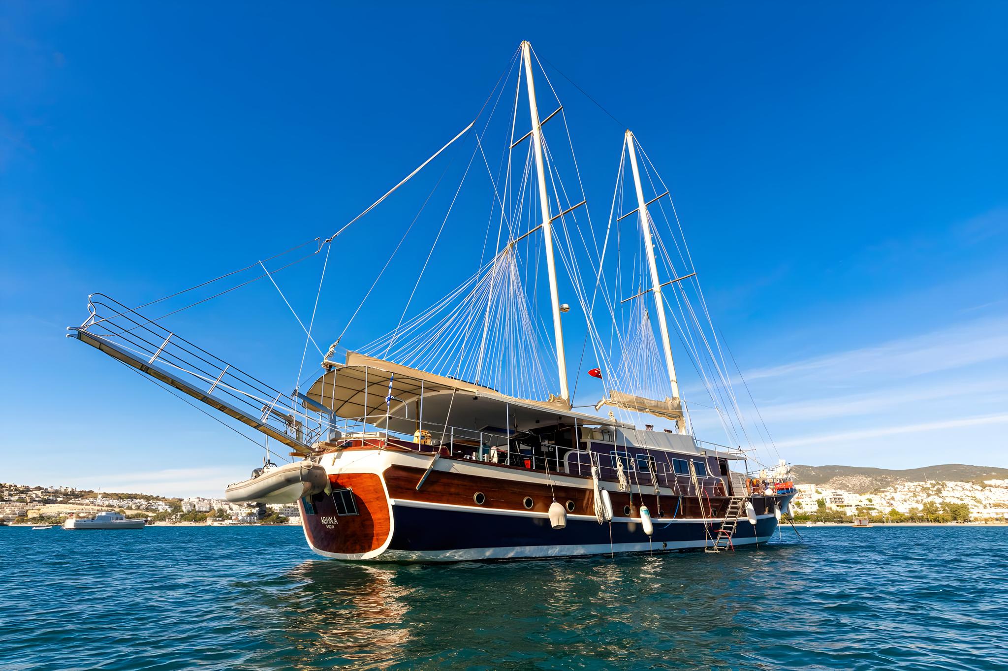 Traditional wooden gulet yacht with three masts anchored in blue Mediterranean waters near coastal town