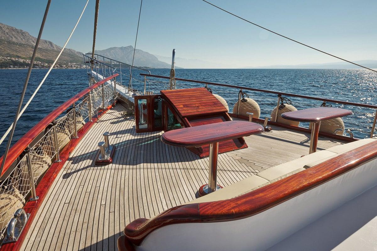 gulet fore deck with teak dining tables, wooden railings and Mediterranean coastline backdrop