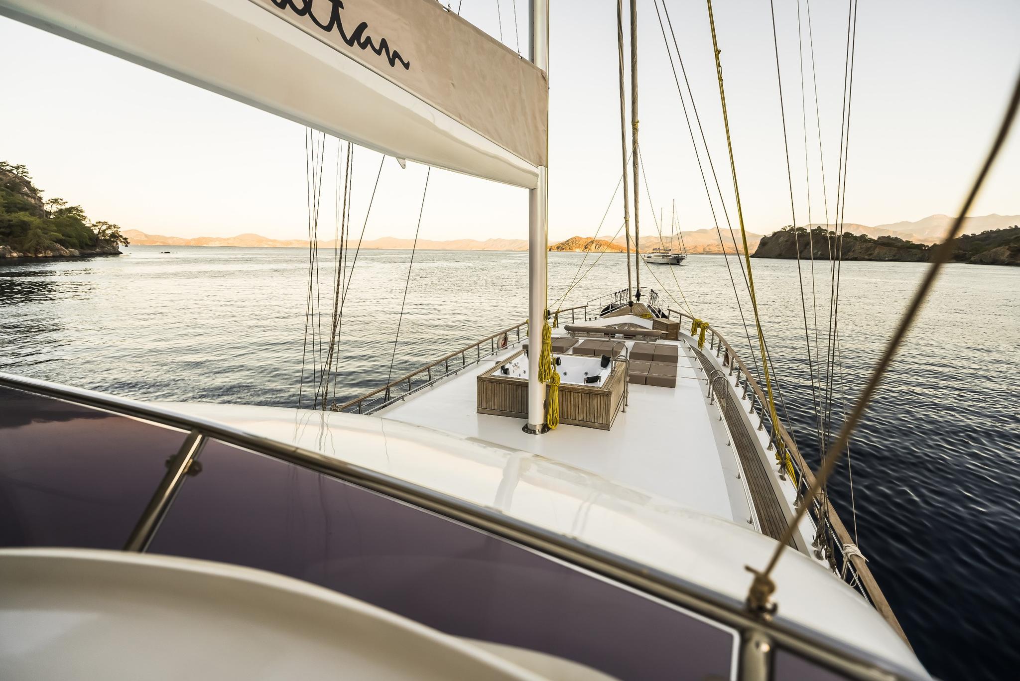 bow deck of sailing yacht with white canvas boom, rigging lines, and coastal bay view at sunset