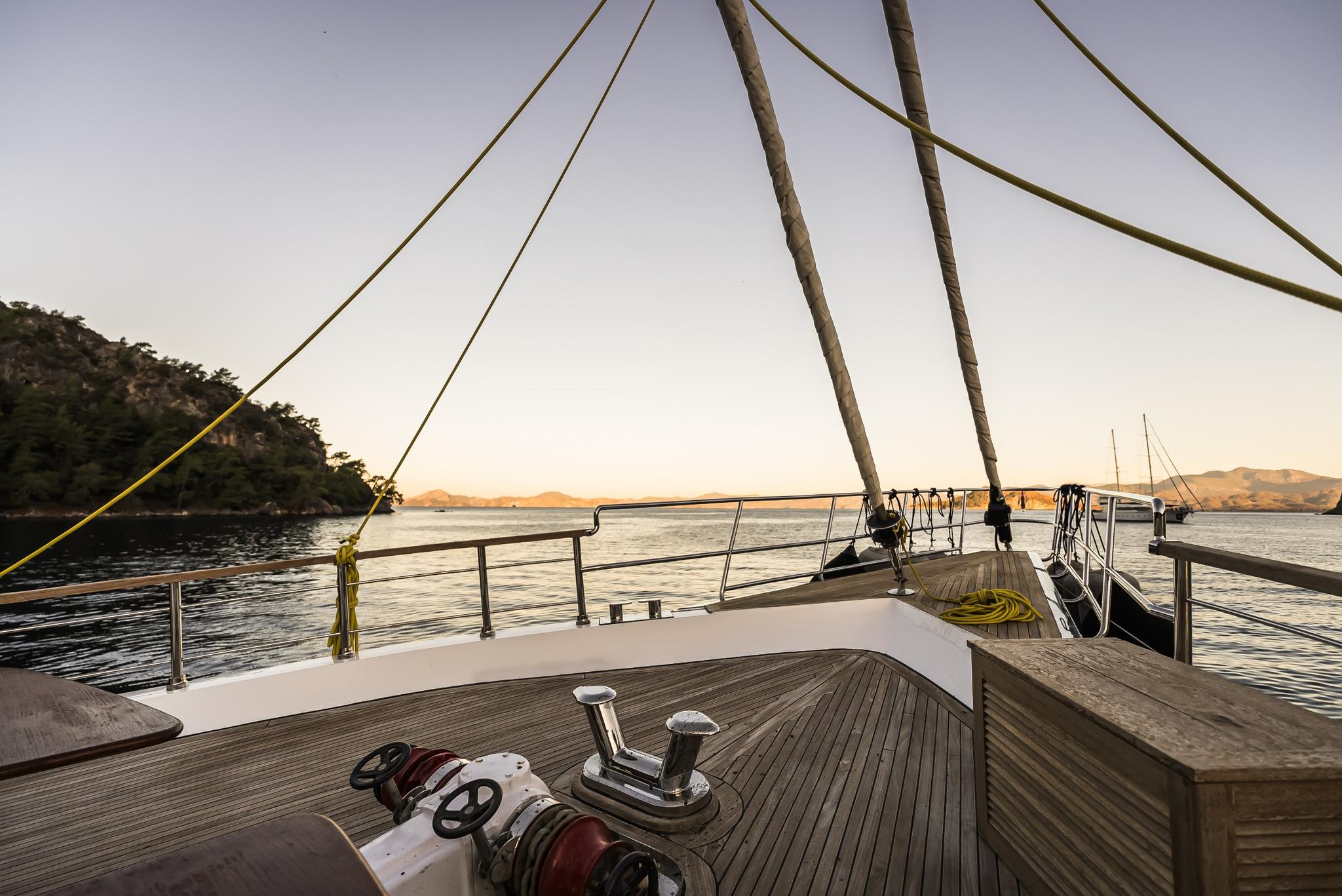 gulet foredeck with wooden deck, rigging lines, windlass and coastline at sunset