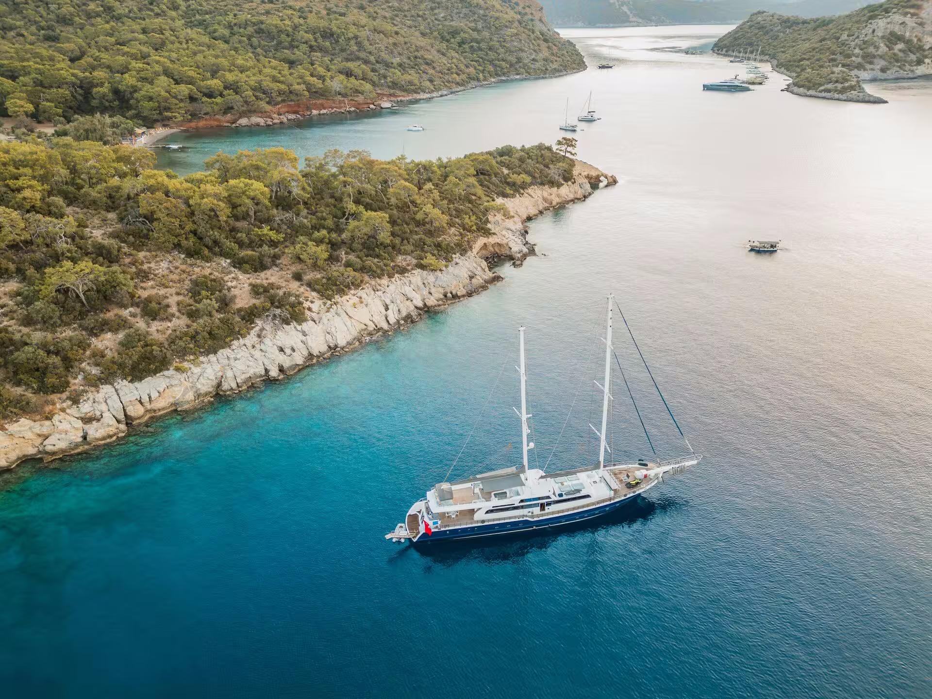 aerial view of white gulet with two masts anchored in crystal clear turquoise bay surrounded by forested hills