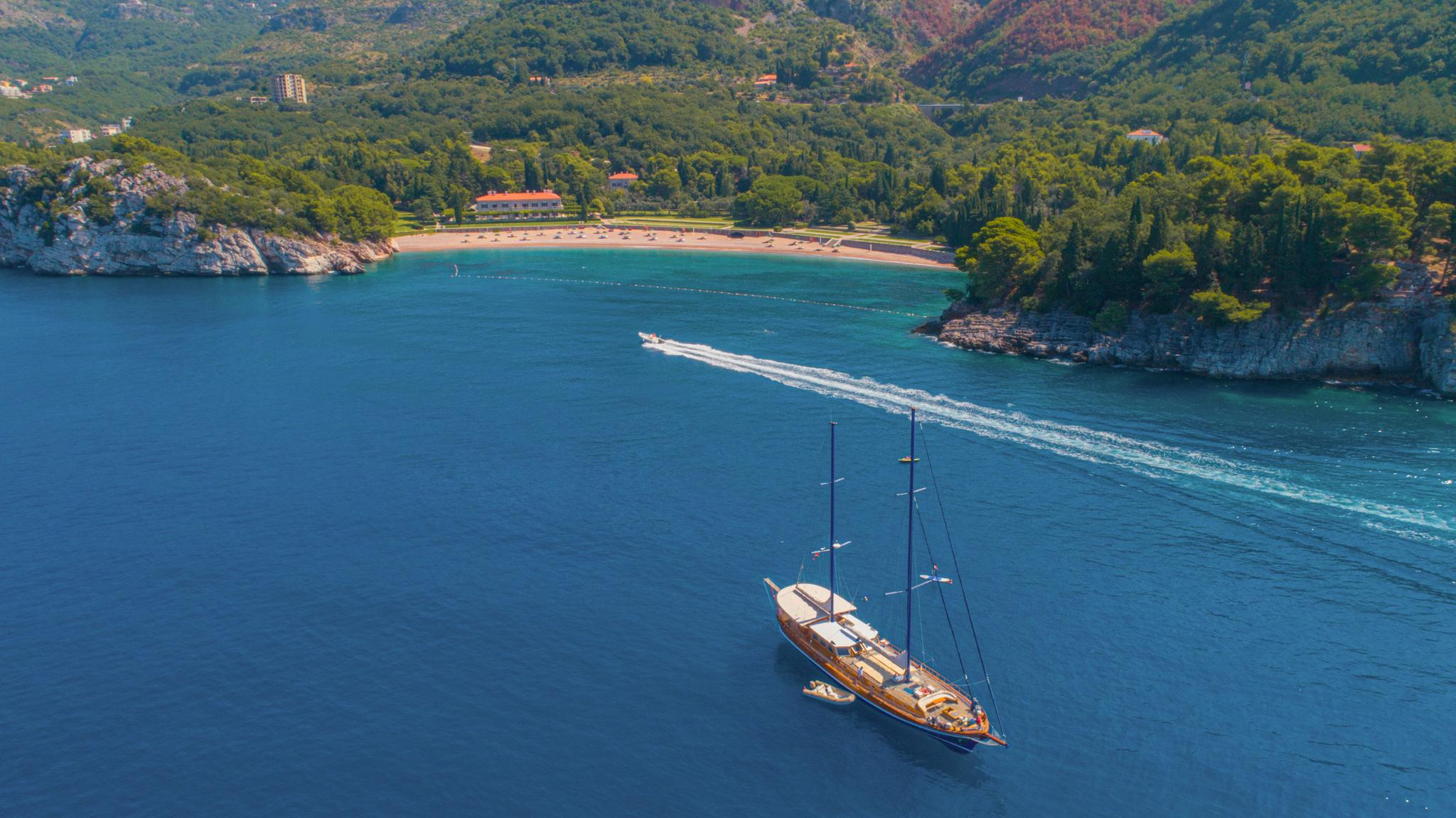 Aerial drone view of classic wooden two-masted gulet anchored in sheltered turquoise bay with sandy beach and forested hillside