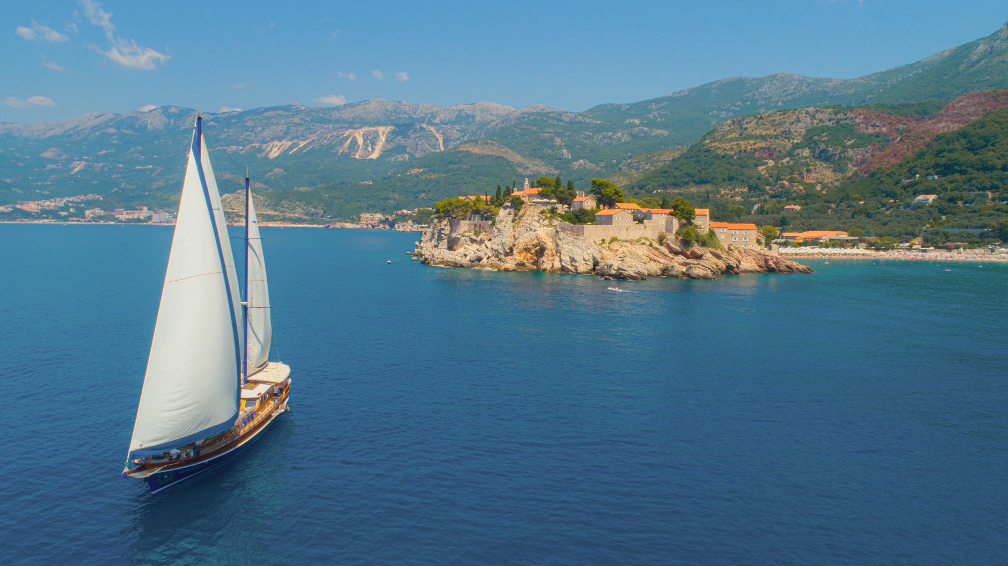 Aerial drone view of wooden gulet sailing under full white sails past a historic rocky island with orange-roofed buildings