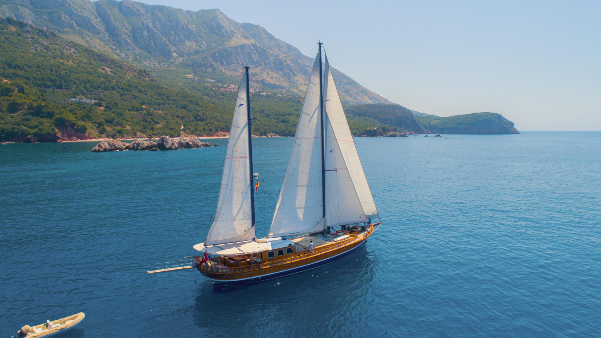 Aerial drone view of a twin-masted wooden gulet sailing with full white sails against a mountainous coastline