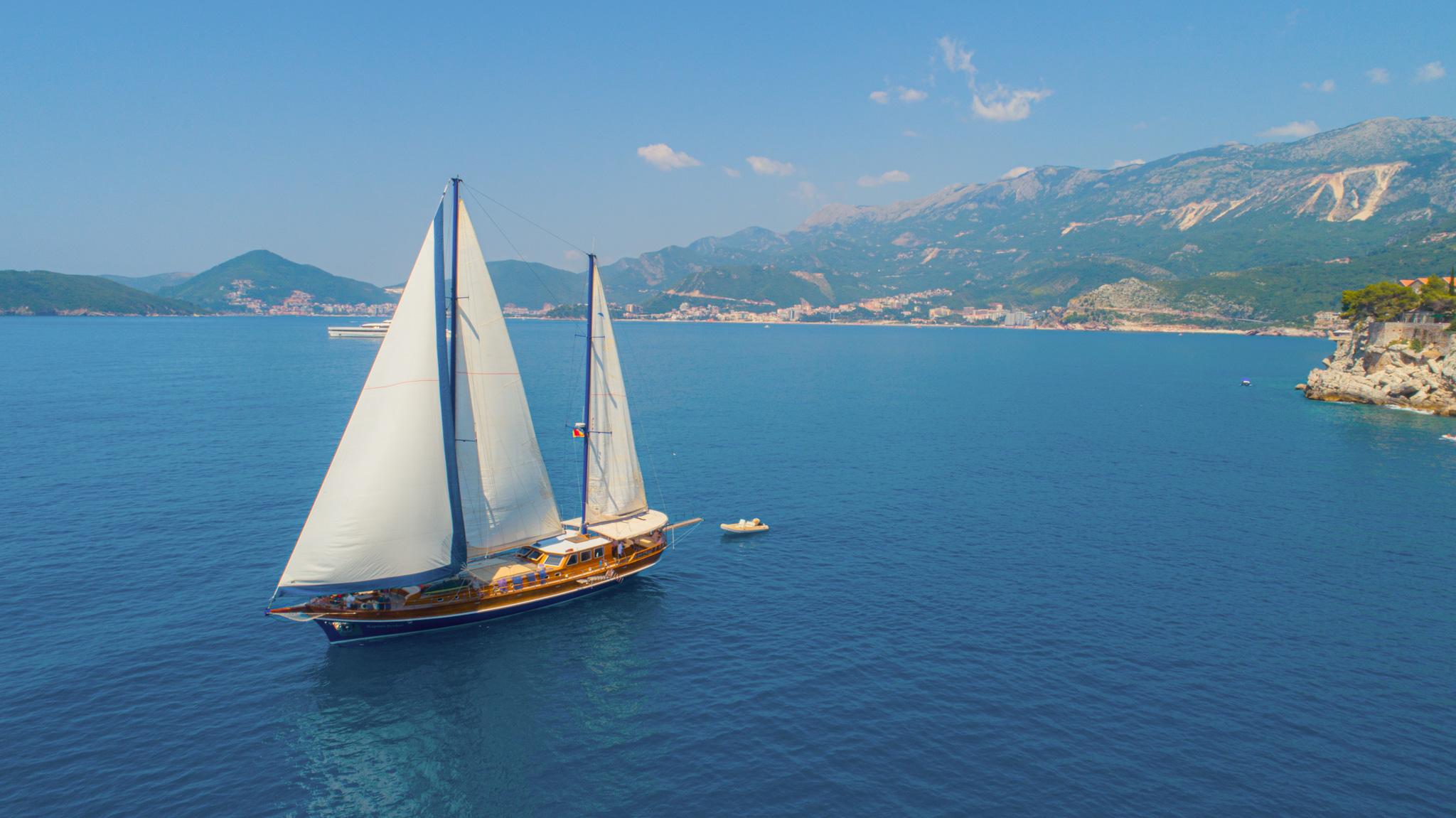 Aerial drone view of a traditional wooden gulet sailing with full white sails near rocky coastline and mountains