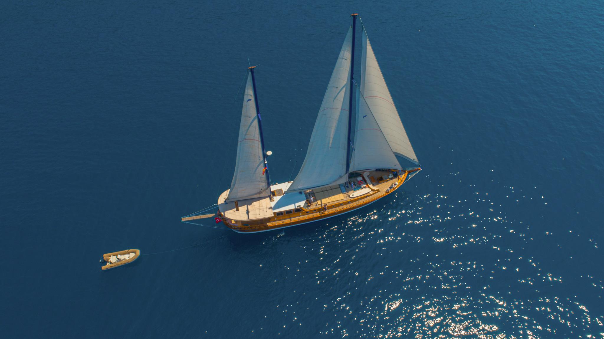 Aerial drone view of wooden gulet sailing under full white sails on deep blue sea with inflatable tender nearby