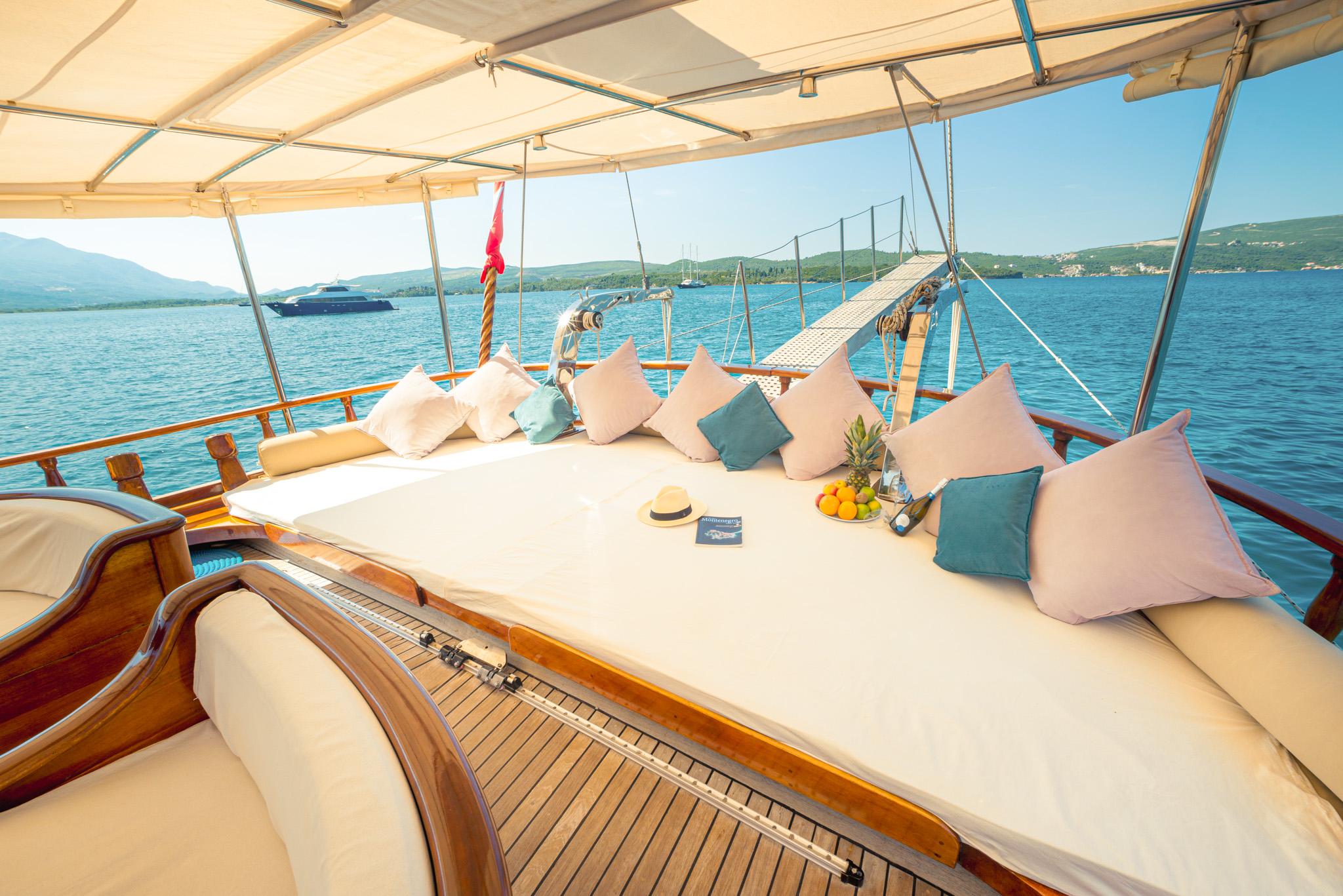 gulet foredeck with large cushioned sun lounger, pink and teal scatter cushions, panama hat, fruit platter and bottle, sheltered under cream canvas awning