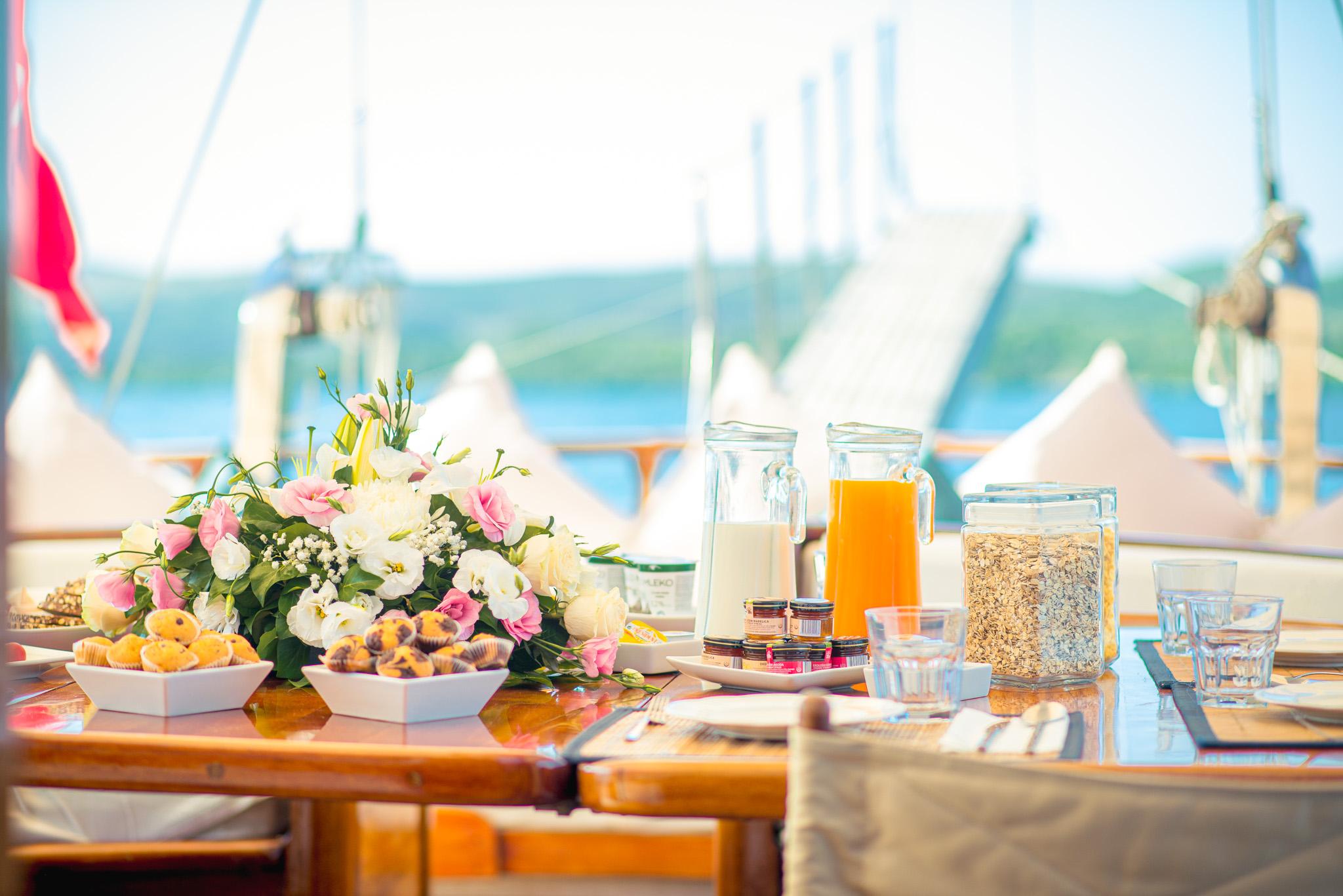Luxury gulet breakfast table with fresh muffins, orange juice, milk, muesli, jam jars and pink and white flower arrangement