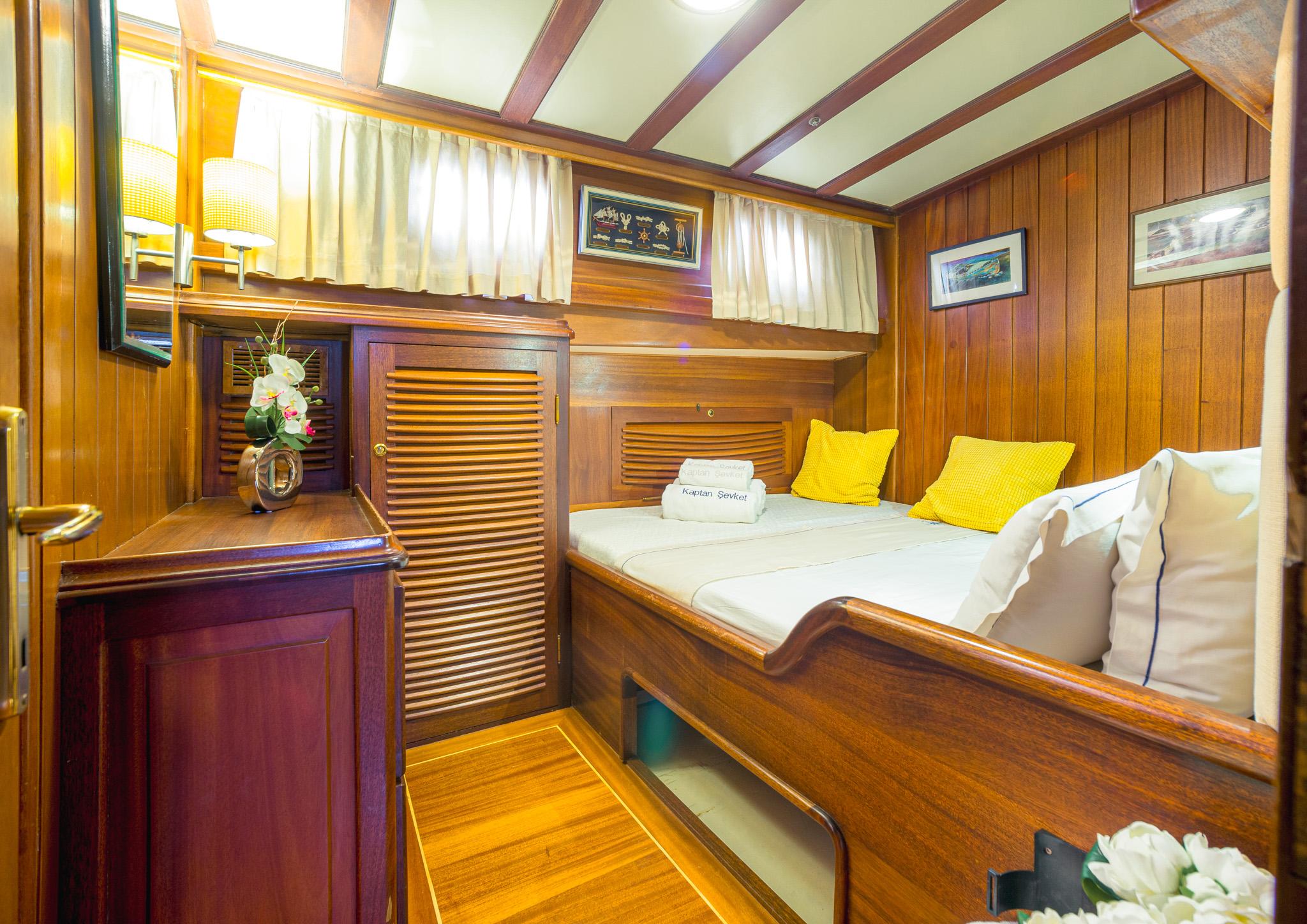 Gulet double cabin with rich teak woodwork, louvred wardrobe doors, white bedding and yellow accent cushions, branded Kaptan Sevket towel on bed