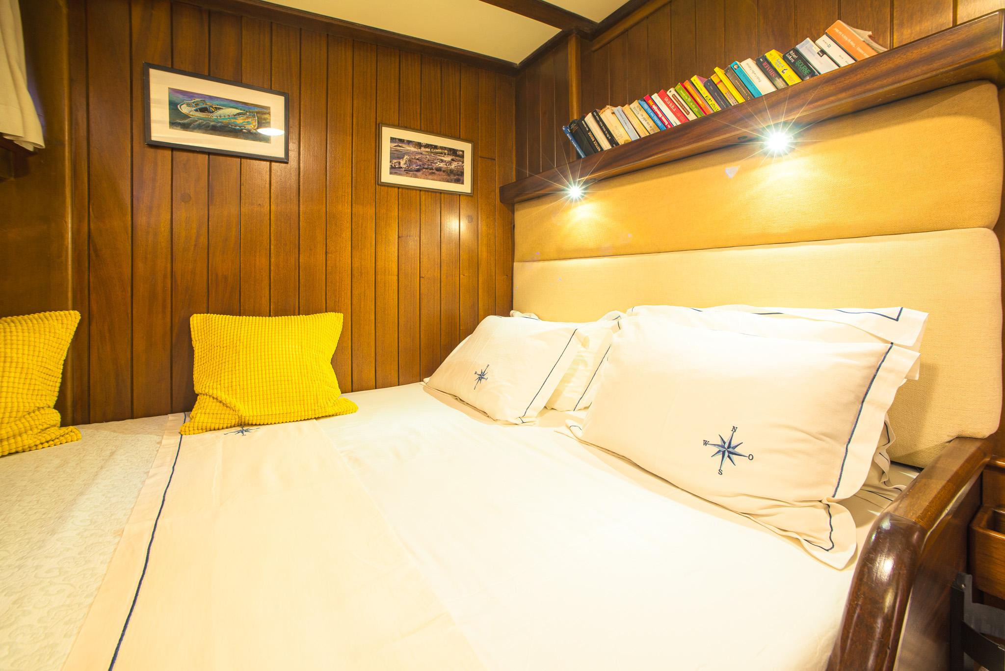 Gulet double cabin with white compass-embroidered bedding, upholstered headboard, reading lights, and built-in book shelf above