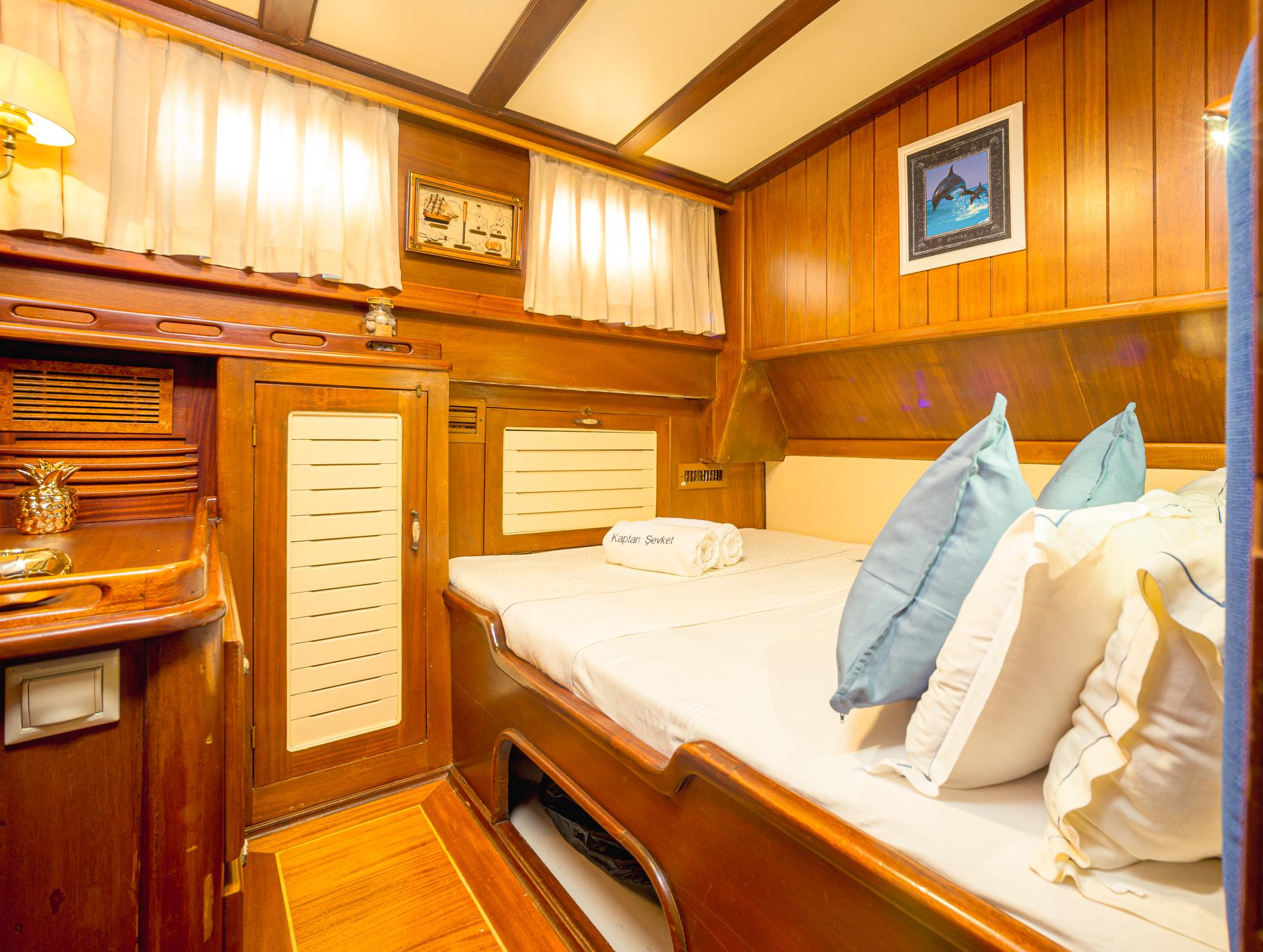 Gulet double cabin with warm teak wood panelling, white bedding, blue cushions, louvred wardrobe doors and porthole windows
