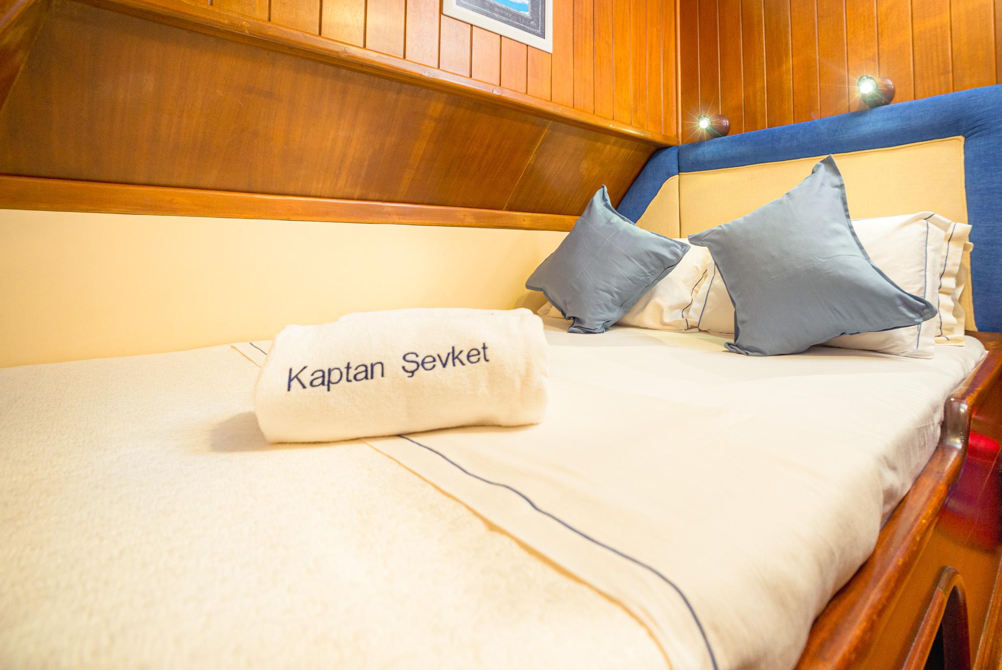Double cabin bed with blue accent pillows and Kaptan Şevket branded towel on gulet