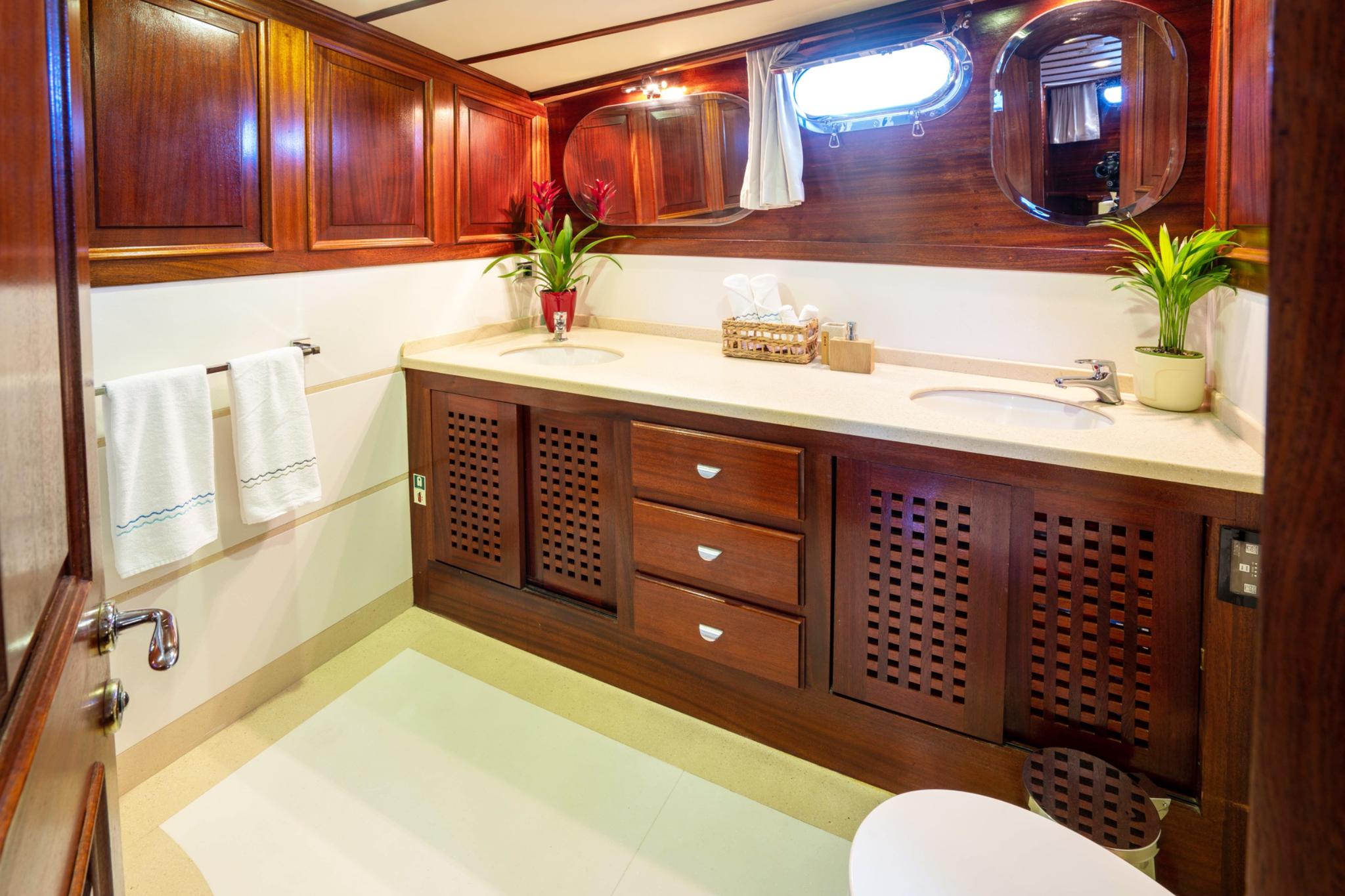 luxury yacht bathroom featuring cream marble bathtub, dual vanity sinks, rich teak cabinetry and oval porthole