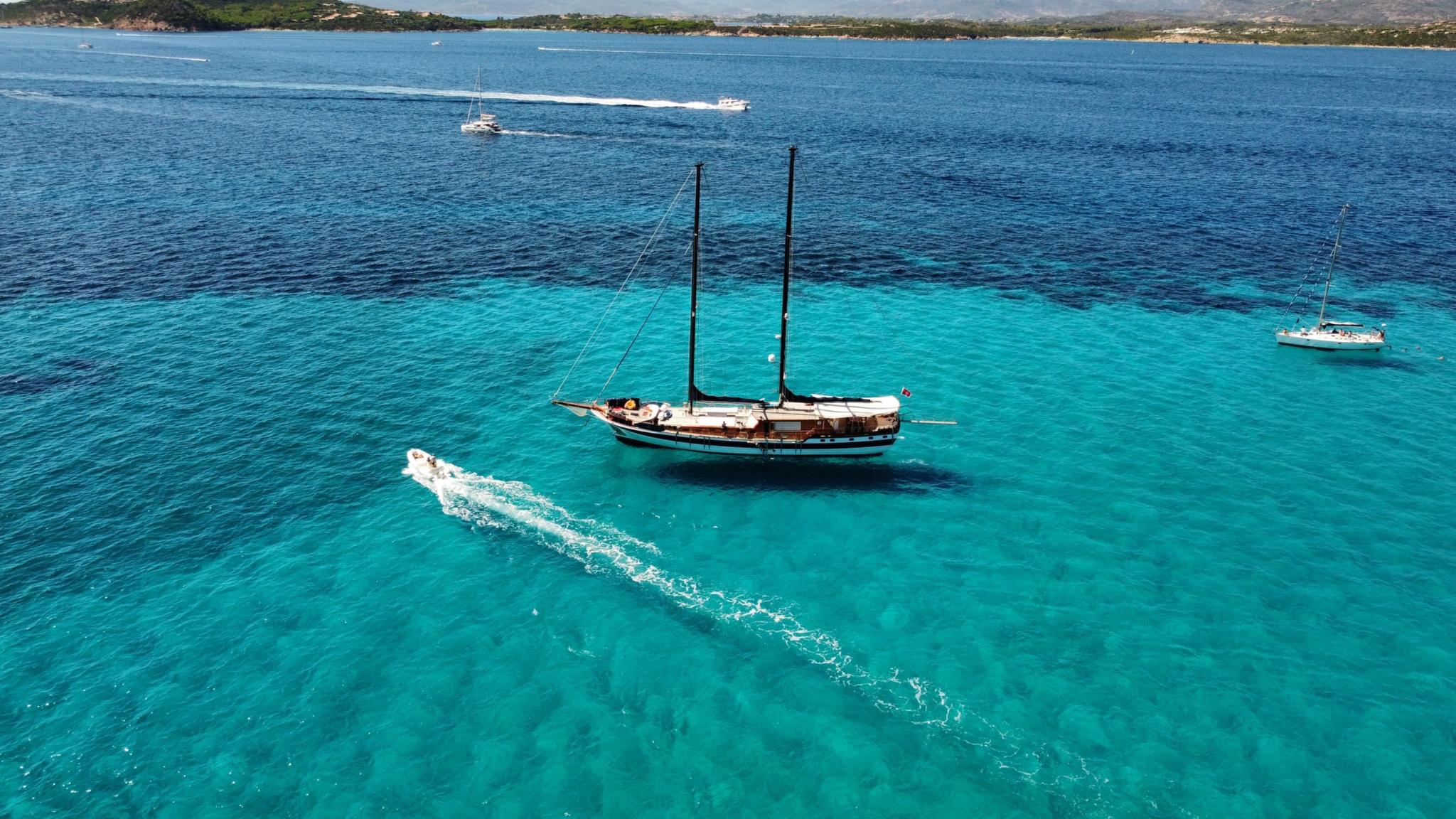aerial view of wooden gulet yacht anchored in crystal clear turquoise waters with white wake trail