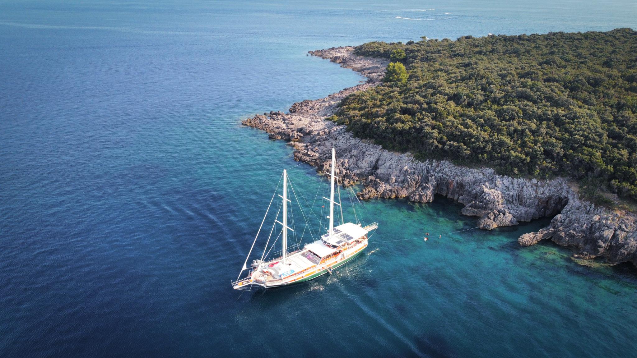 Aerial drone view of a classic gulet anchored in a secluded turquoise cove beside a tree-covered rocky coastline