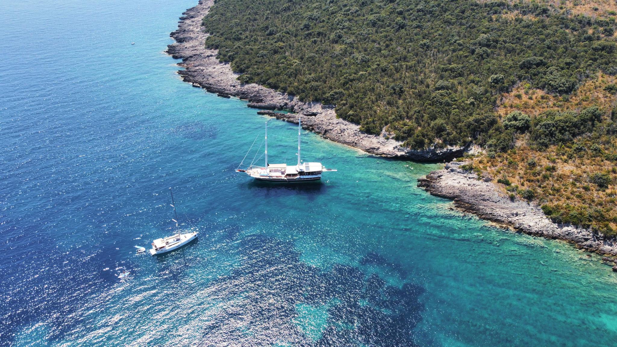 Aerial drone view of a gulet anchored in a clear turquoise bay beside a rocky tree-covered coastline