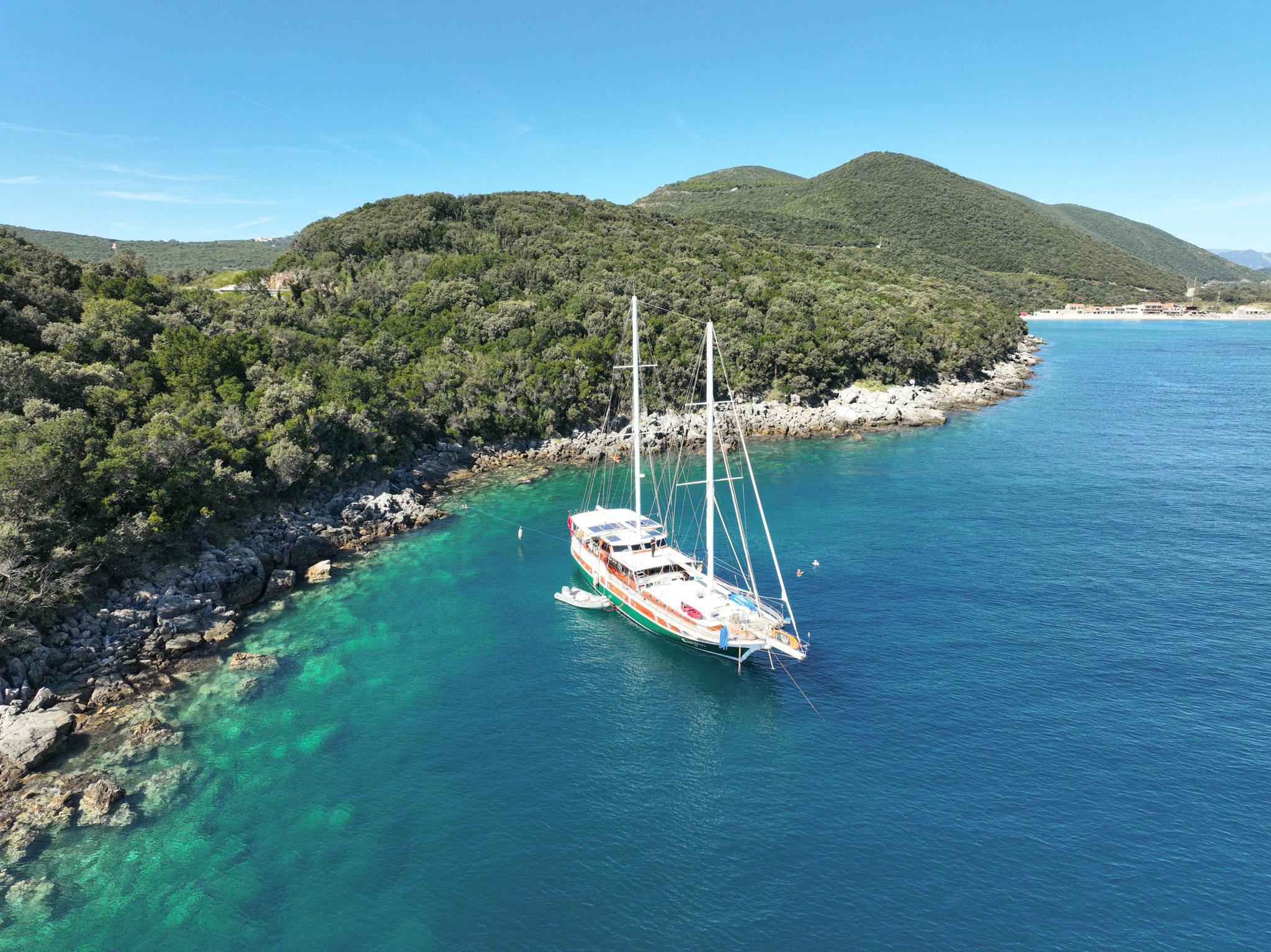 aerial drone view of classic gulet anchored in crystal-clear turquoise bay with forested hills