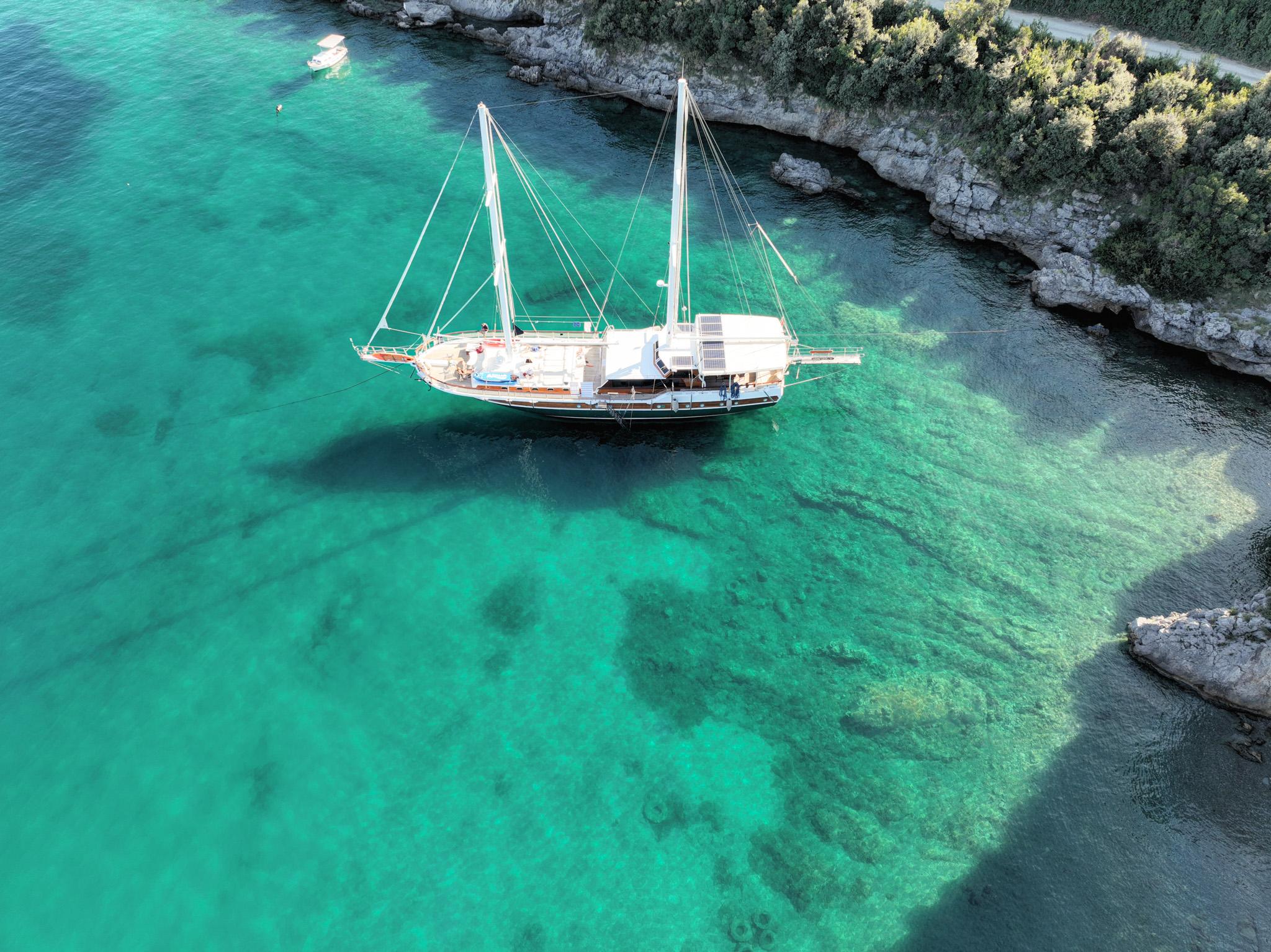 Aerial drone view of a two-masted gulet anchored in a sheltered turquoise bay with rocky pine-covered shoreline