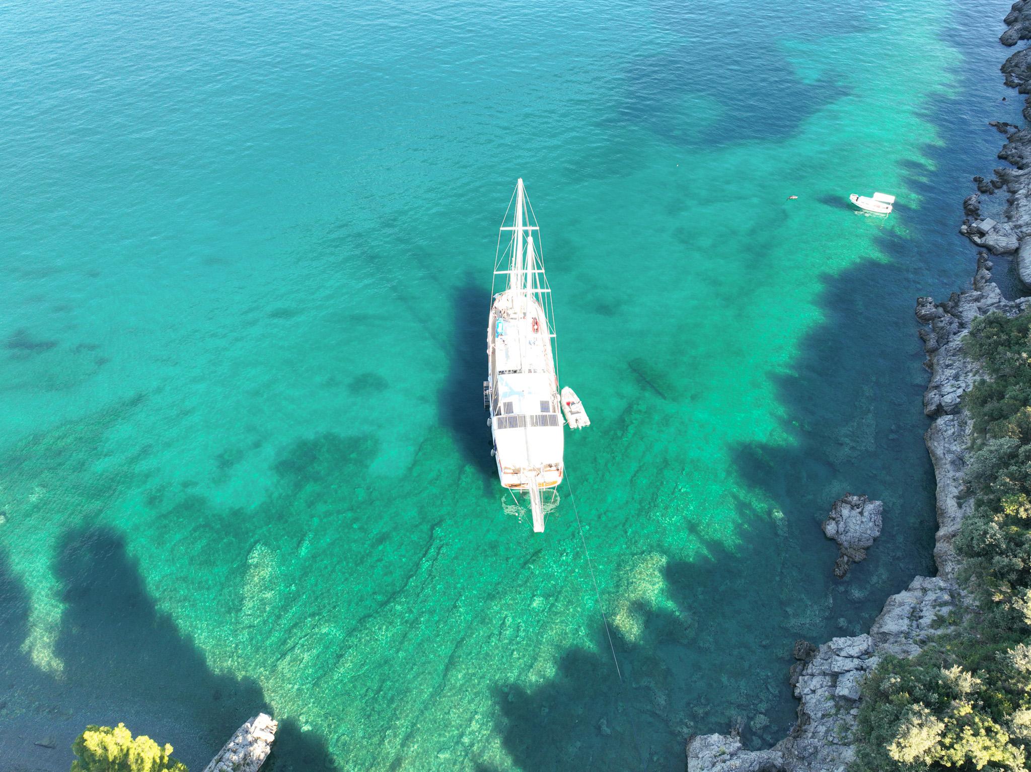 aerial drone view of white gulet anchored in turquoise rocky cove with tender alongside