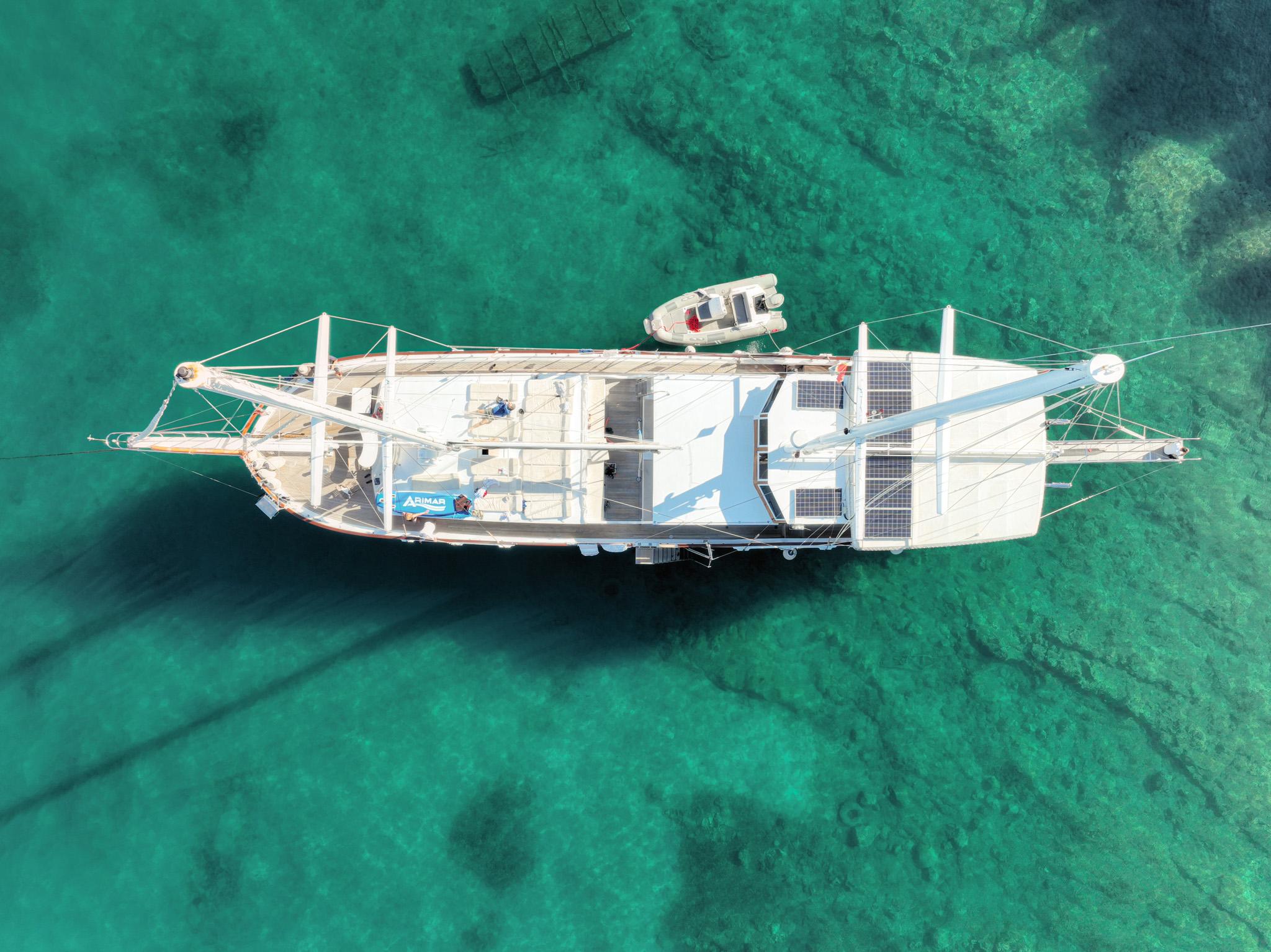 Aerial drone view of a gulet with solar panels and tender dinghy anchored in crystal turquoise waters