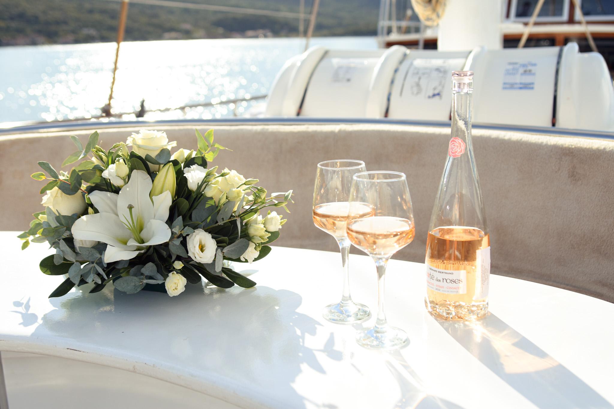 Cote des Roses rosé wine bottle with two filled glasses and white lily bouquet on gulet deck