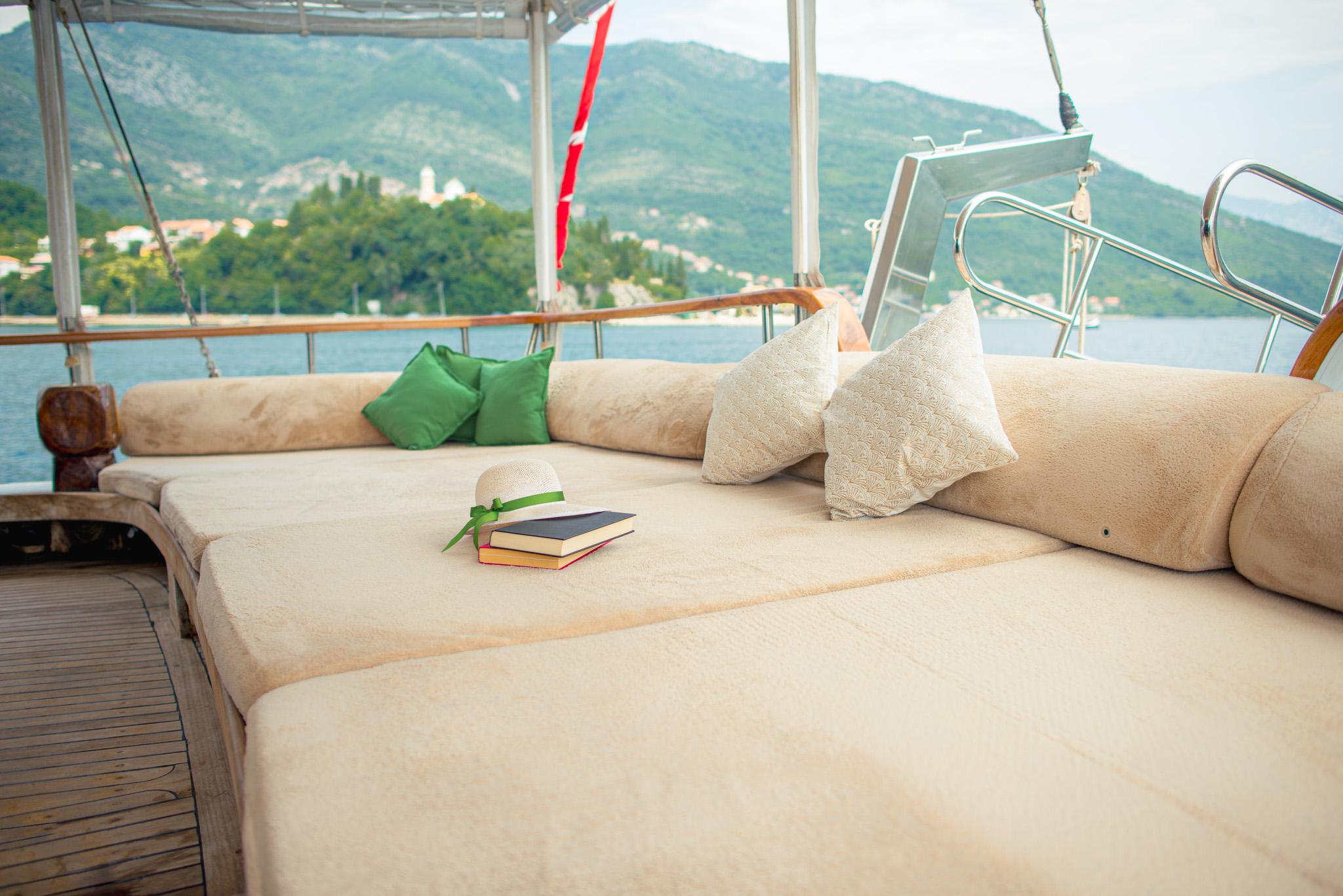 Gulet aft deck wide cream cushioned seating with green and ivory pillows, two stacked books and straw hat, mountainous coastline backdrop