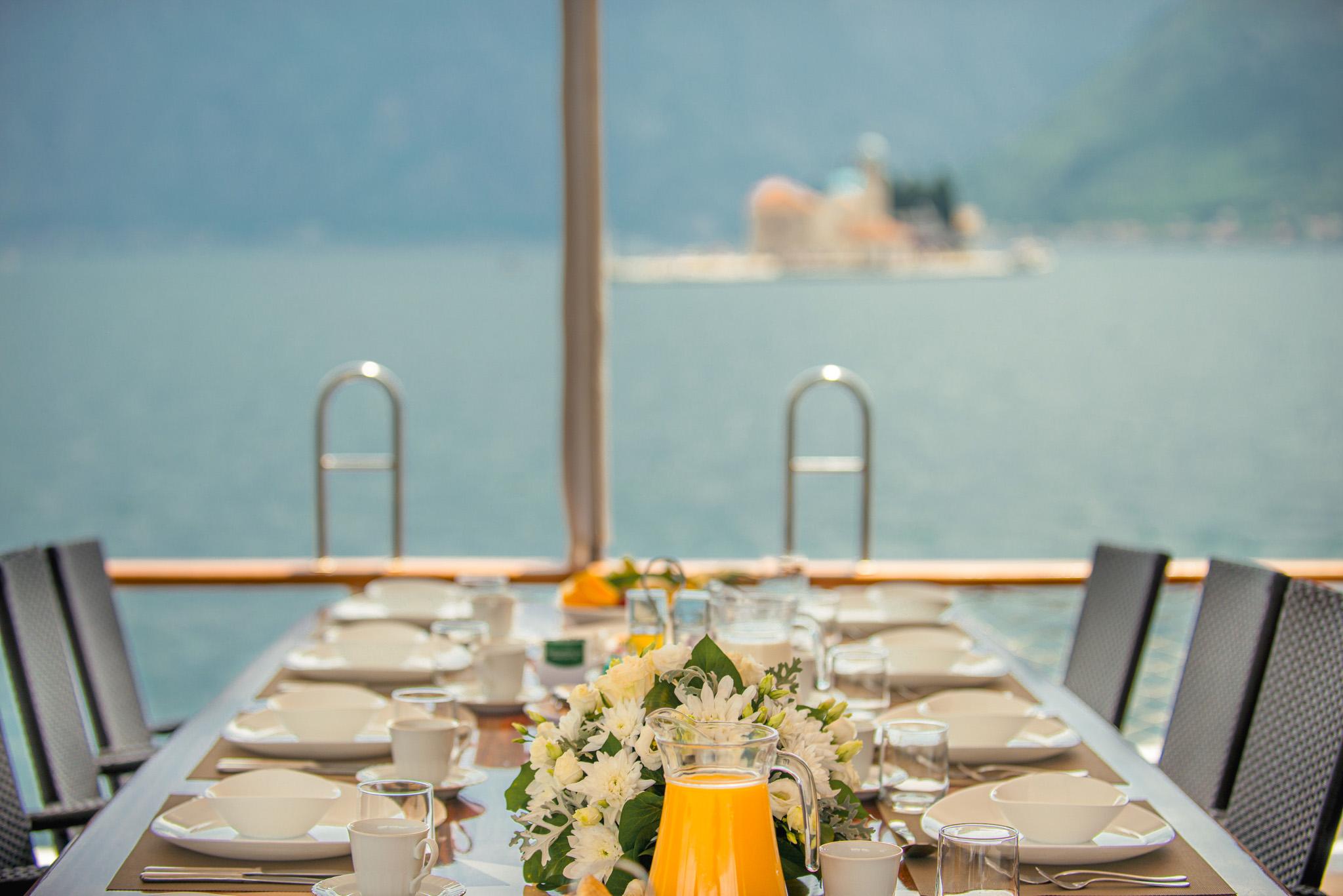 Luxury gulet outdoor dining table set with white crockery, fresh white flowers, and a pitcher of orange juice, with a small island church visible across the bay