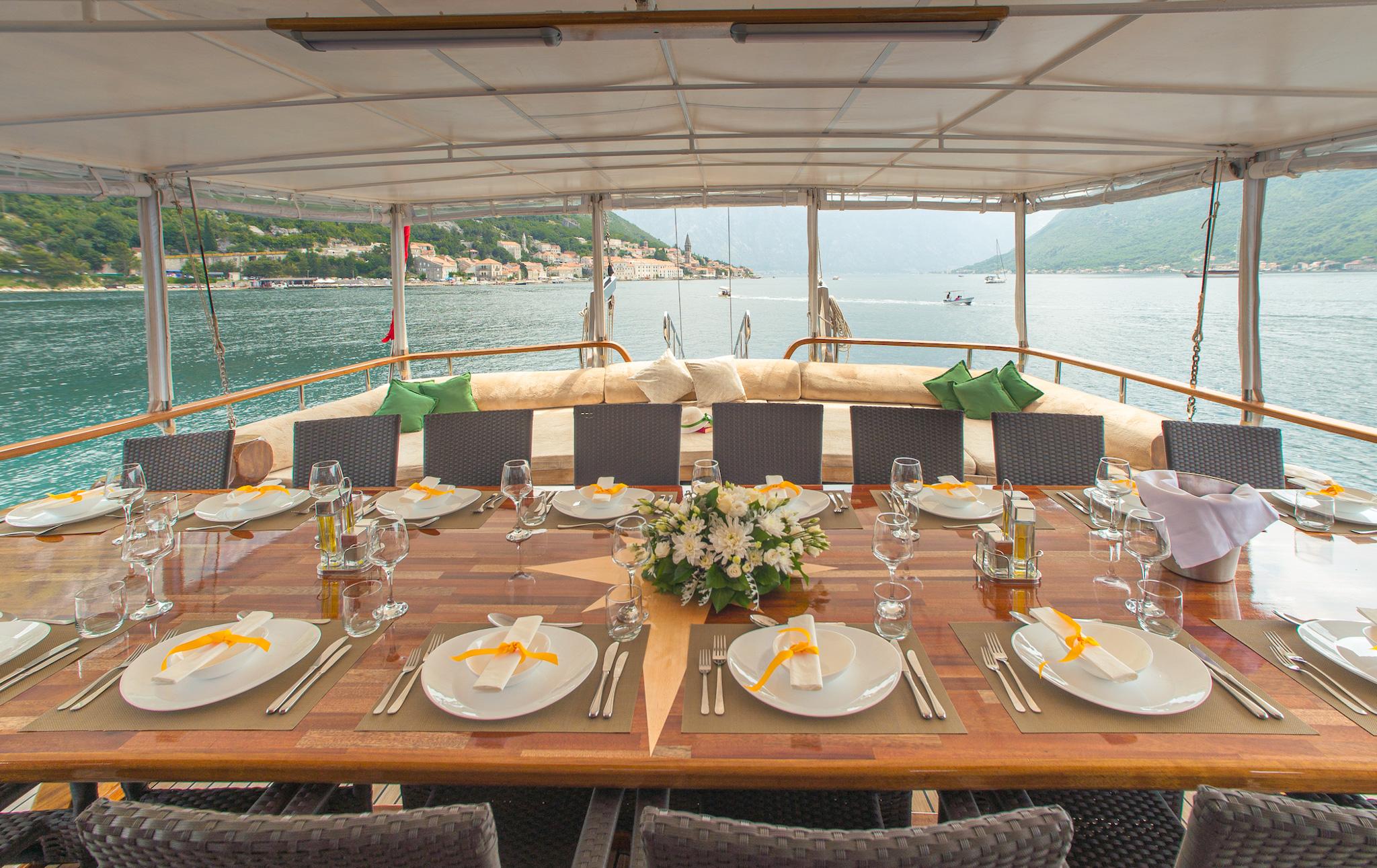 Elegantly set outdoor dining table on gulet deck with white plates, wine glasses, yellow napkins, and white floral centrepiece, overlooking a Mediterranean coastal town