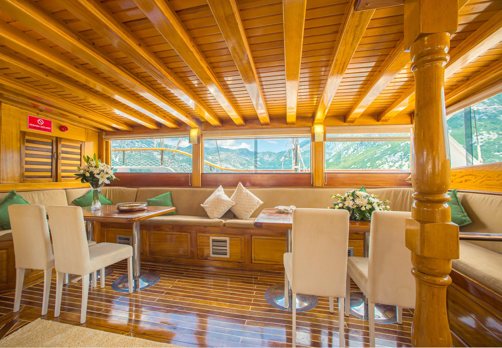 Gulet indoor saloon with varnished wood beamed ceiling, cream upholstered bench seating, white dining chairs, and panoramic windows overlooking mountainous coastline