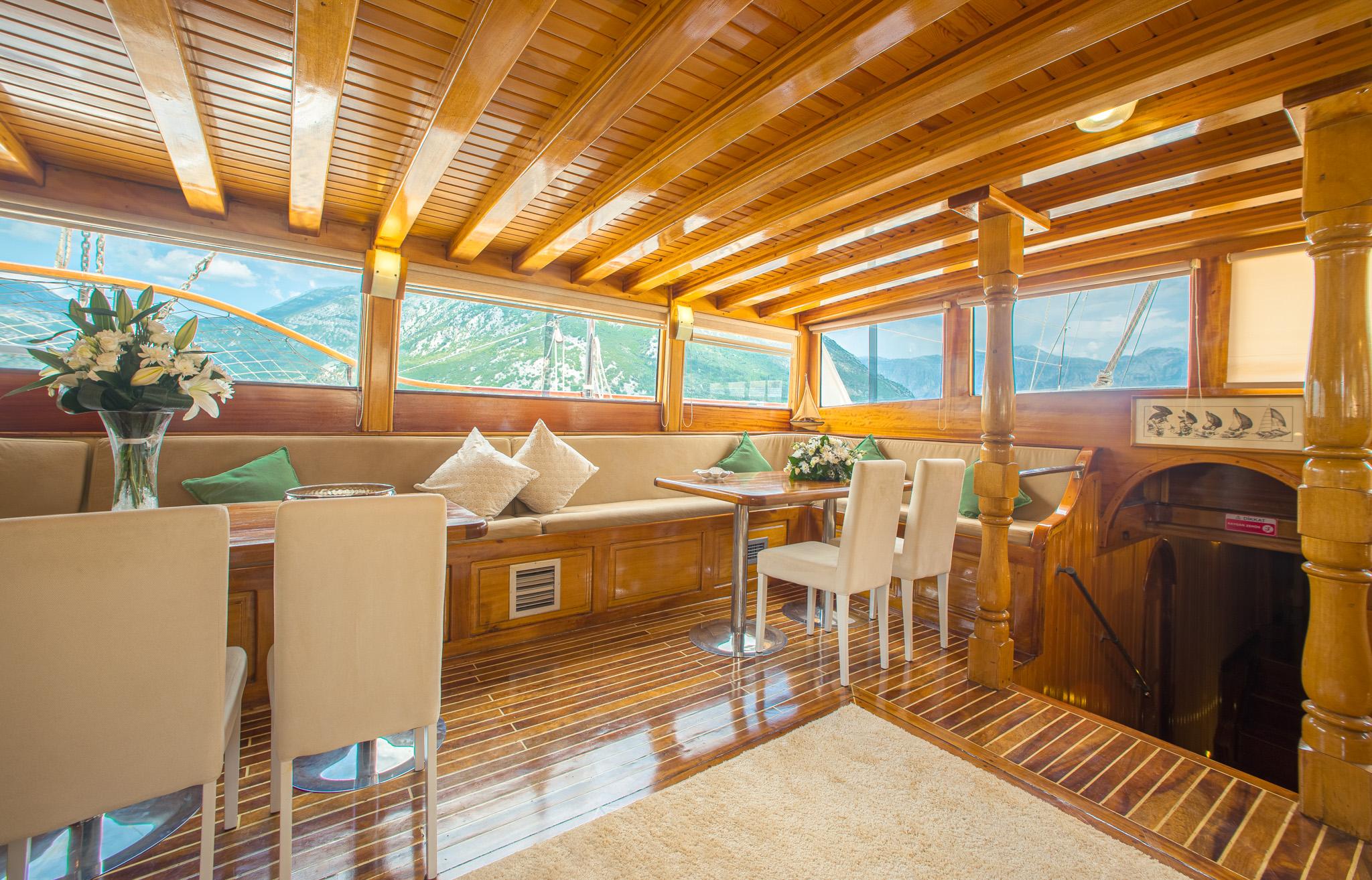 Gulet interior saloon with teak ceiling beams, cream upholstered bench seating, white dining chairs, and panoramic windows overlooking mountains