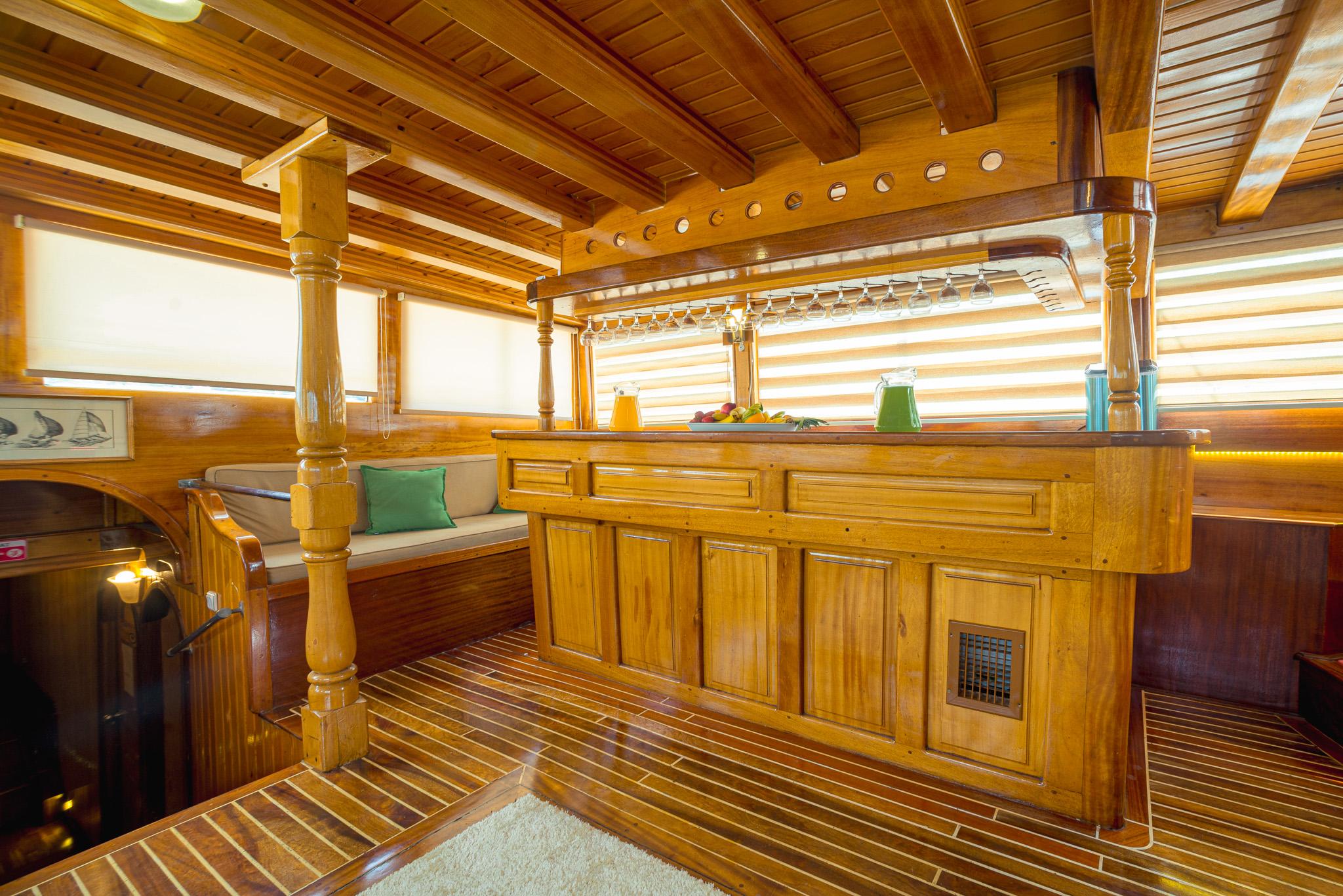 Gulet saloon interior featuring a solid teak bar counter with suspended wine glass rack and fresh juice pitchers