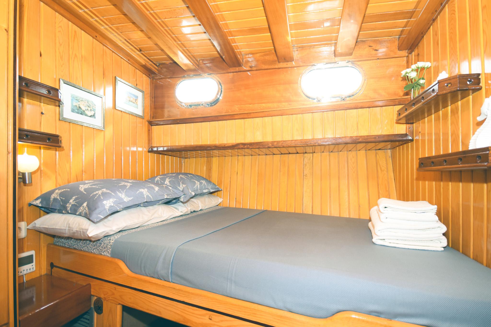 Gulet double cabin with warm wood panelling, two oval portholes, grey bedding and folded towels